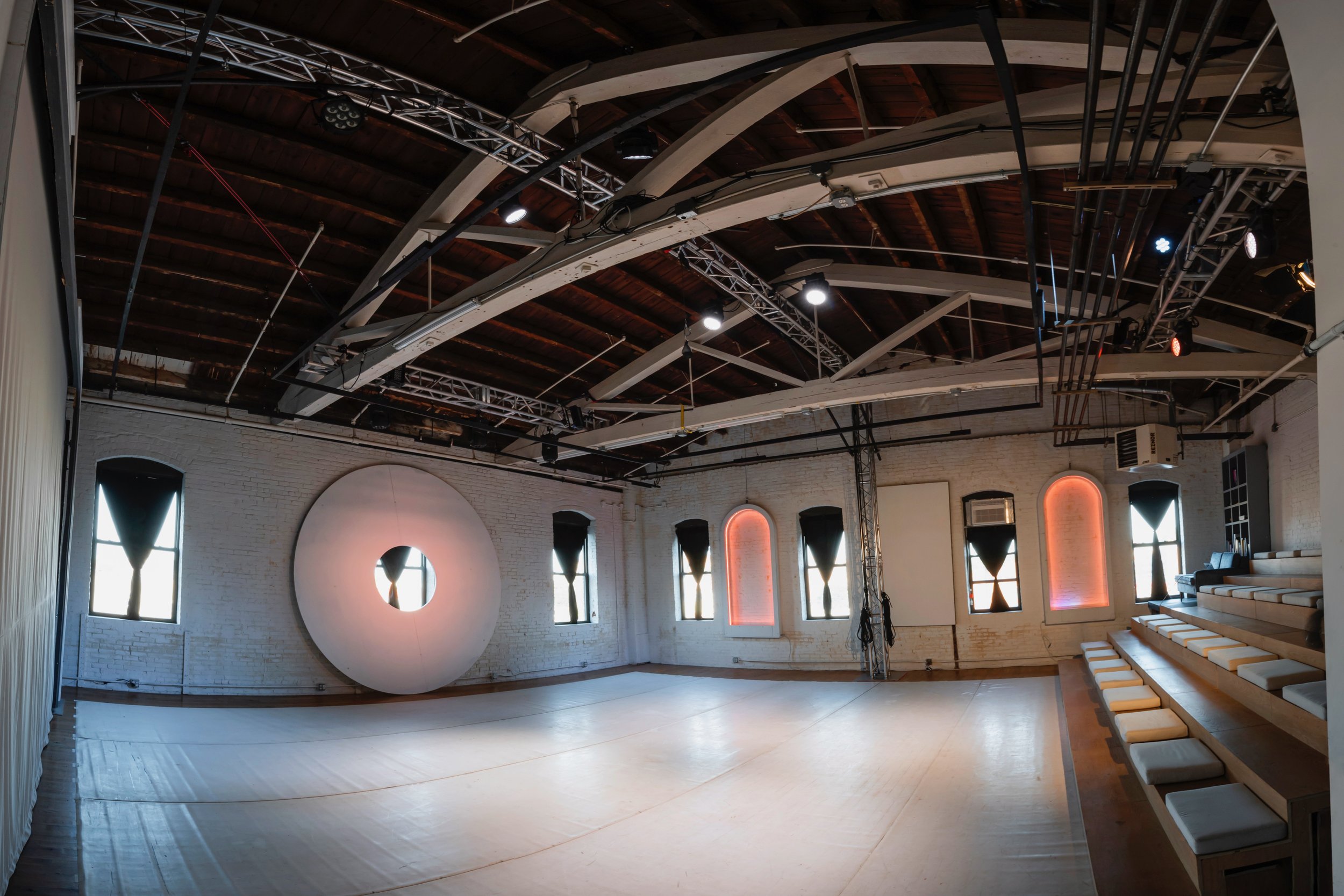 Empty event or performance space with a polished wood floor, white brick walls, and arched windows with black curtains. A large circular art piece with a glowing center is on one wall, and there are tiered seating with white cushions along one side. The ceiling has exposed wooden beams, lighting, and technical equipment.