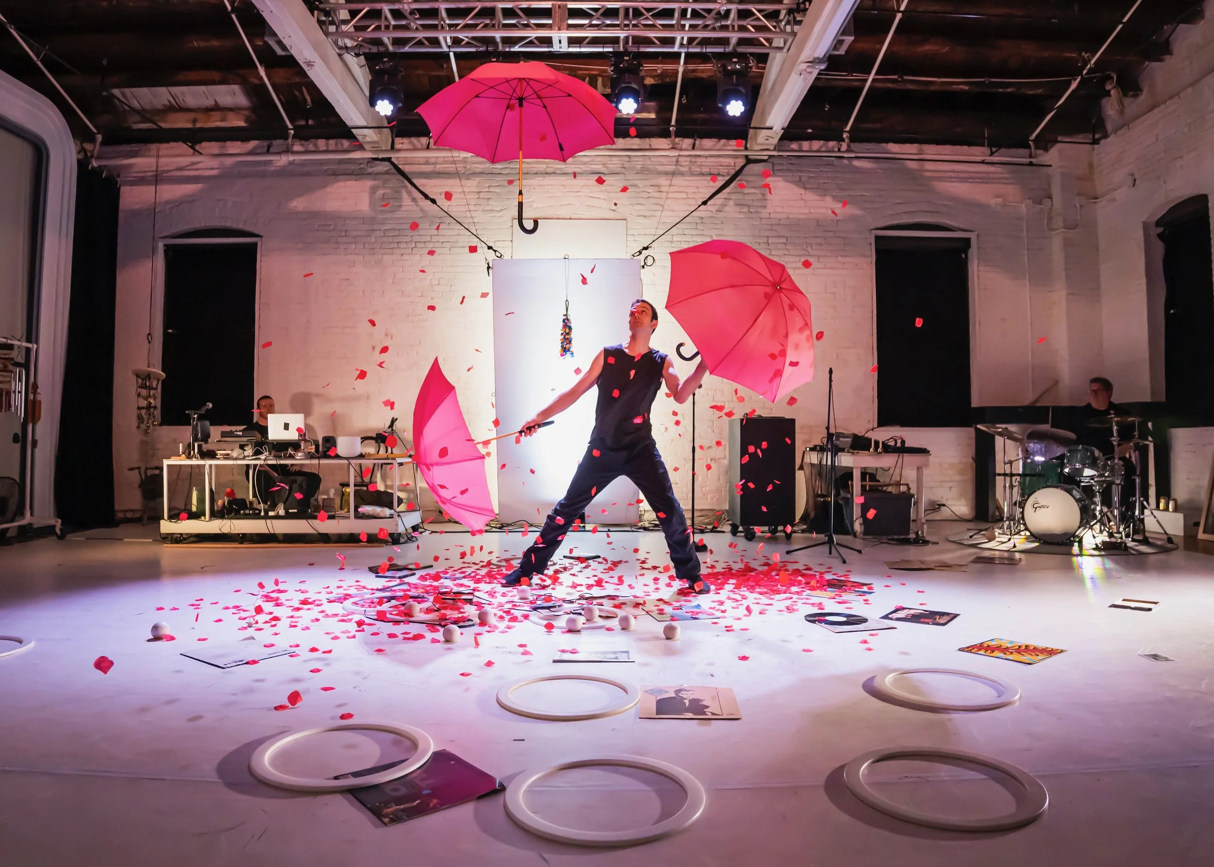 Performer with three pink umbrellas on stage with red confetti, scattered papers, and hoops on the floor, with music equipment and a drummer in the background.