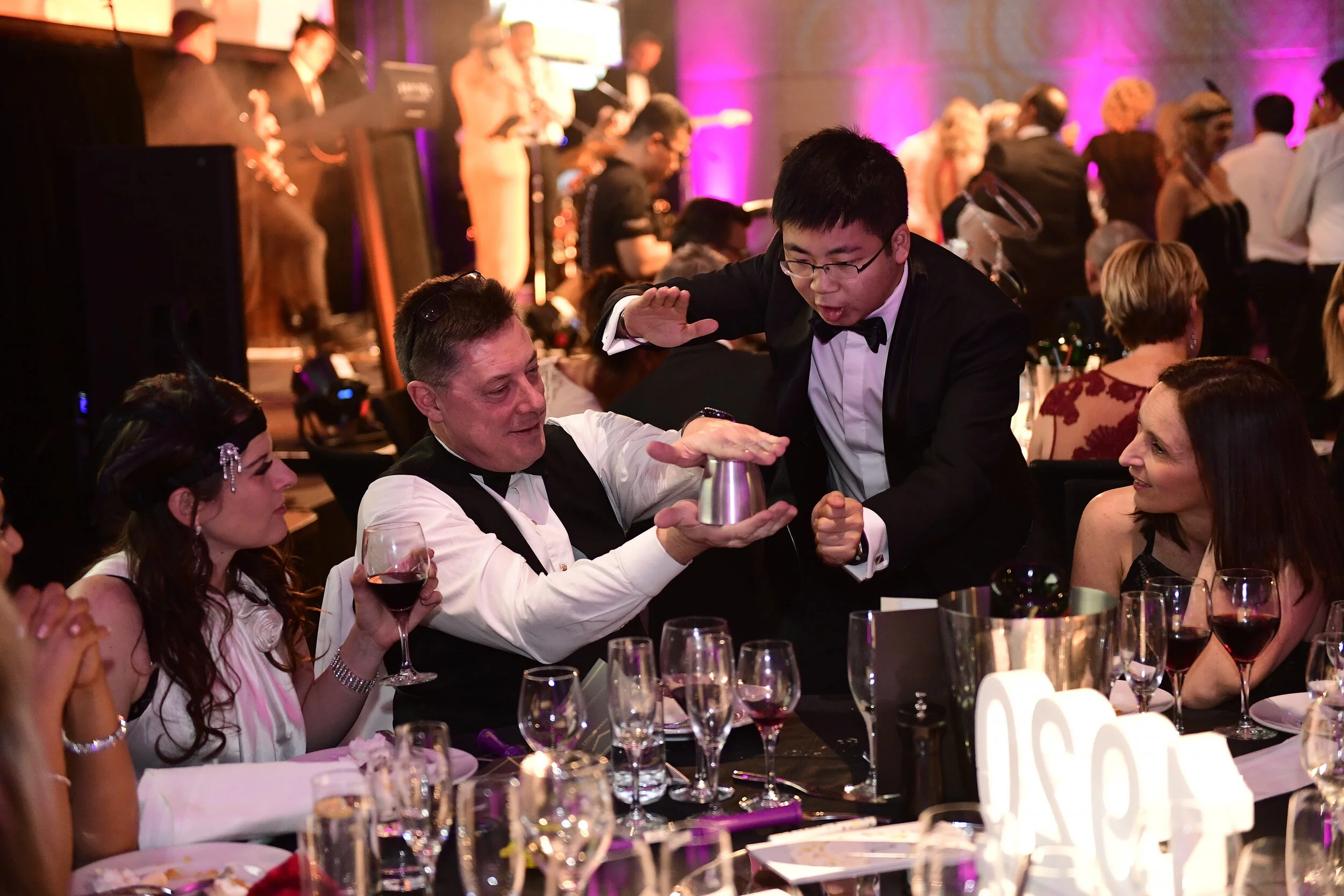 A magician performing a card trick at a formal event, with guests watching at a decorated banquet table.