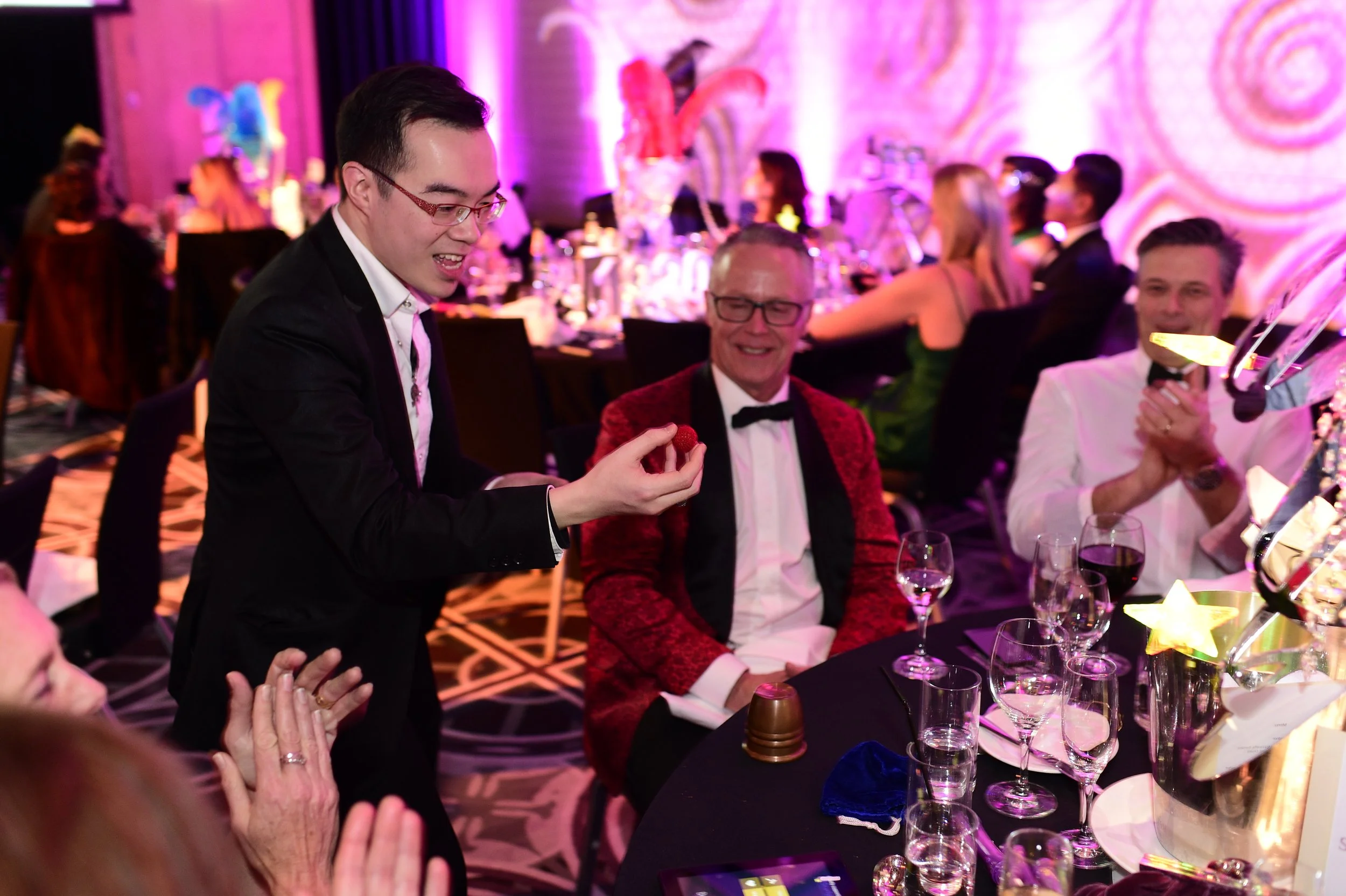 Group of people celebrating with a sydney magician at a wedding reception, with one man being lifted and others smiling and laughing, in a decorated banquet hall.