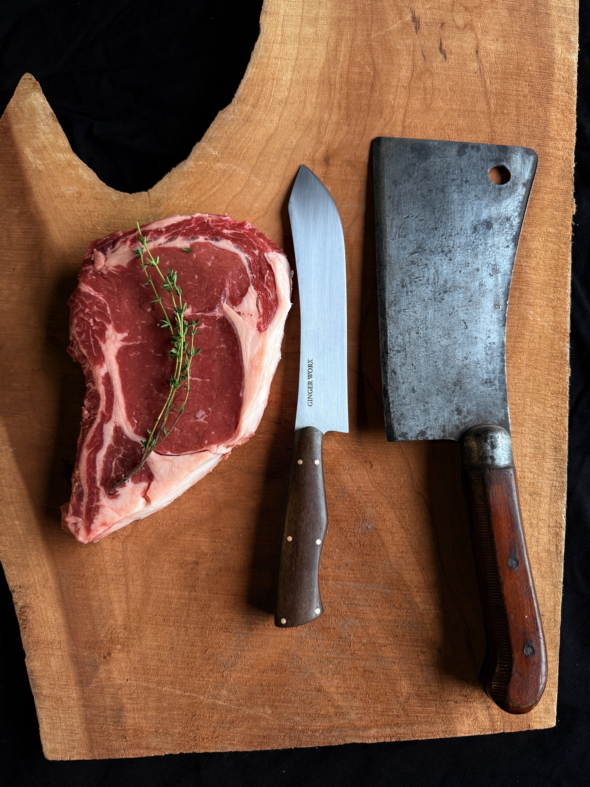 A raw beef steak with a sprig of thyme on top, placed on a wooden cutting board with a kitchen knife and meat cleaver.