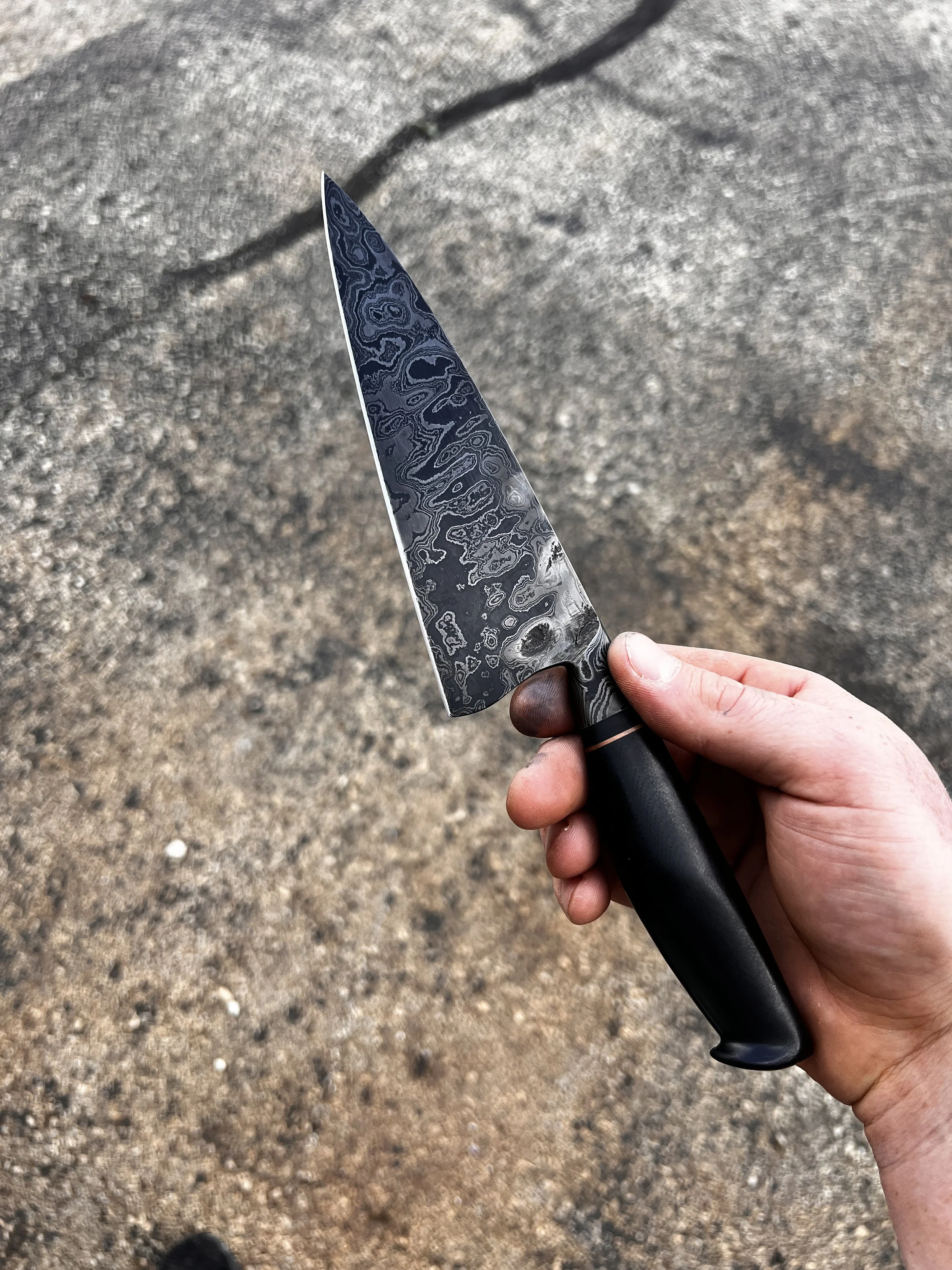 A hand holding a Damascus steel kitchen knife with a black handle over a black top