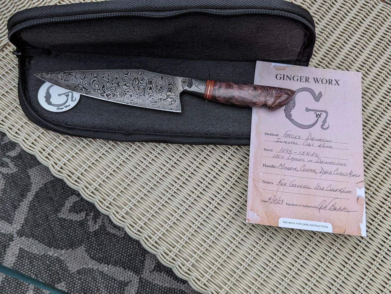 A Damascus steel chef knife with a wood and copper handle resting inside a black zippered case, placed on a woven rattan table next to a handwritten note and a sticker.