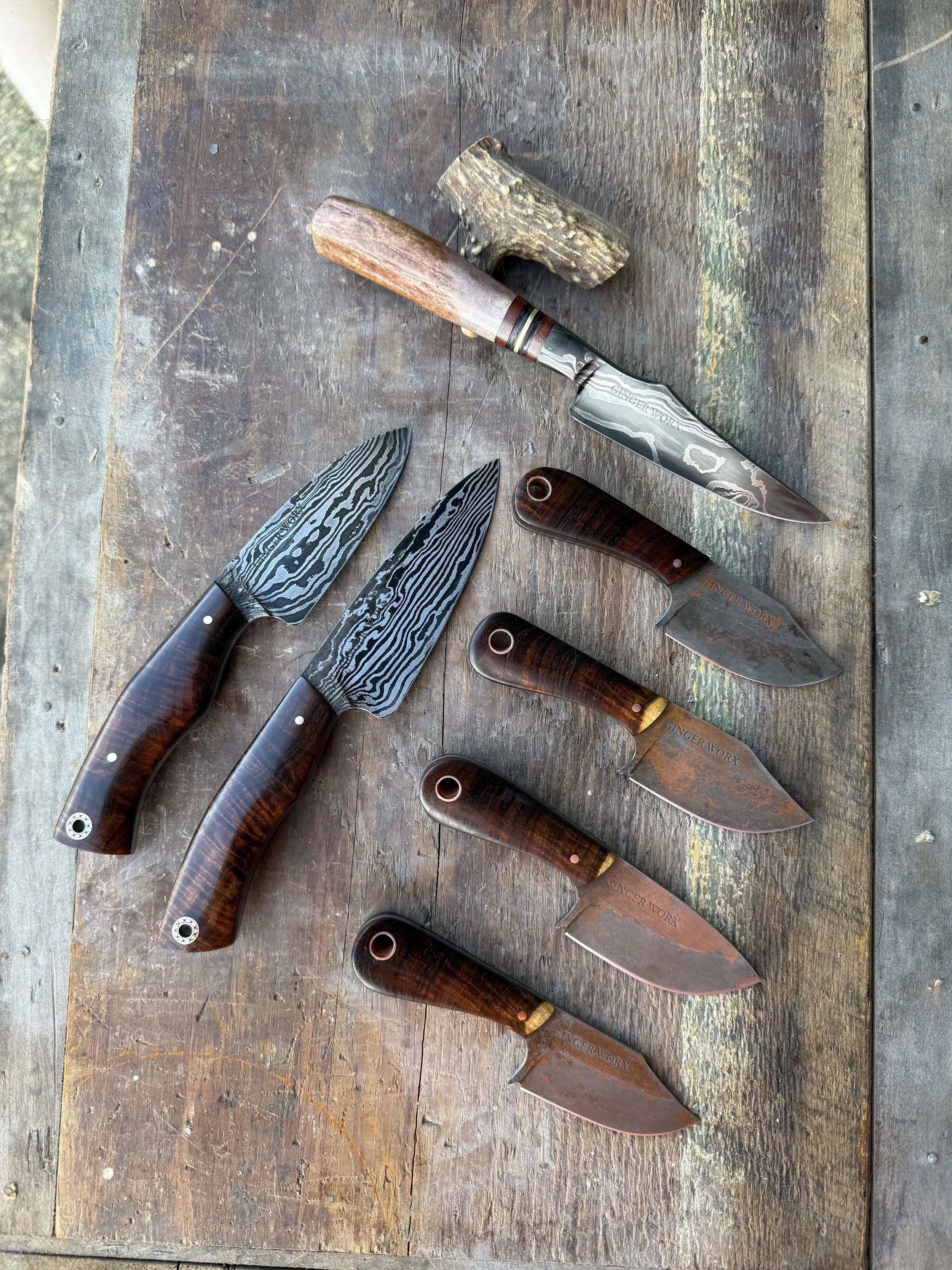 Six handcrafted knives with wooden handles, etched blades, and rustic designs, arranged on a weathered wooden surface with a piece of wood acting as a stand.