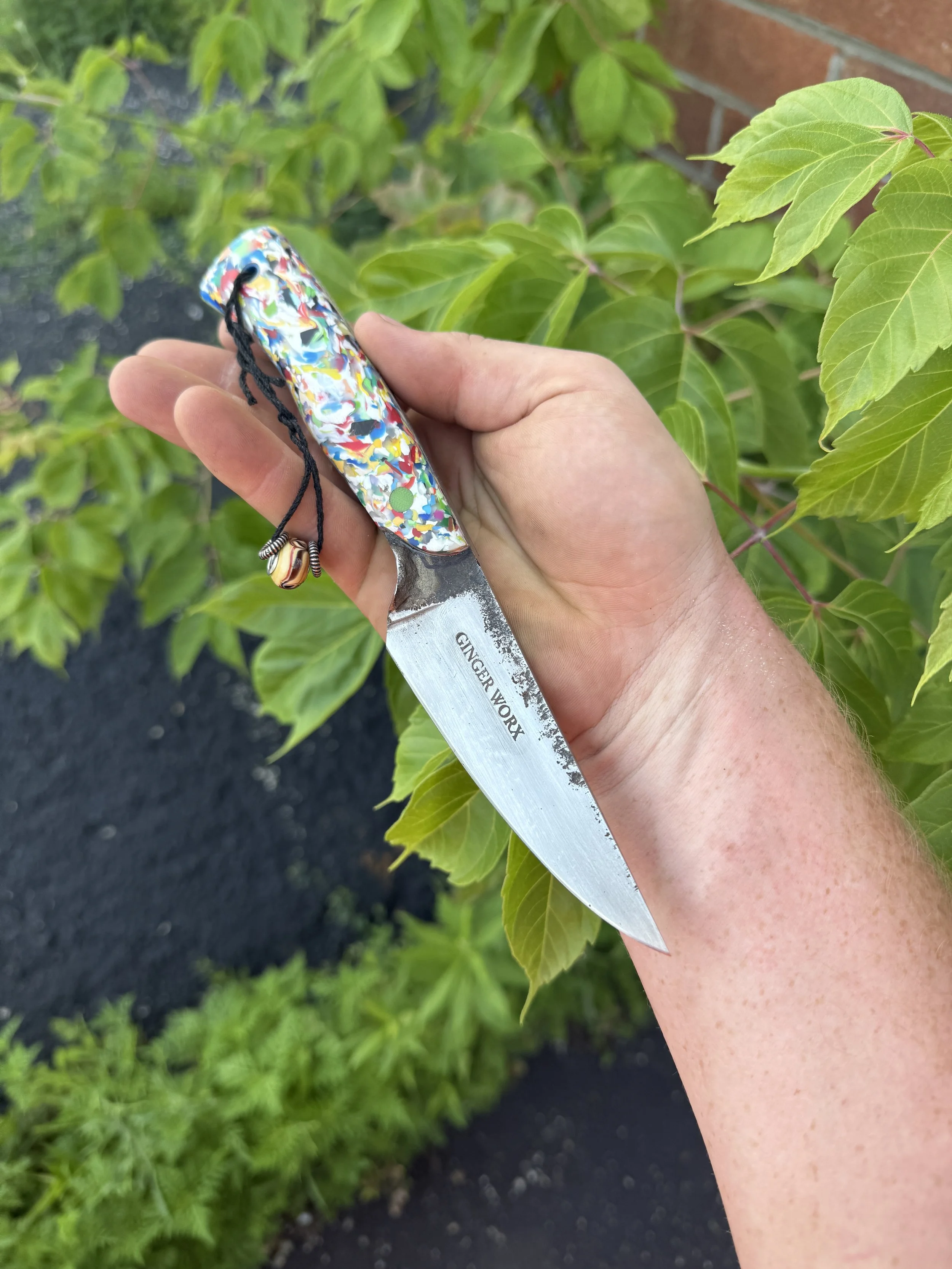 A hand holding a colorful, patterned pocketknife with a black cord and a small decorative bead attached, against a backdrop of green plants and a brick wall.