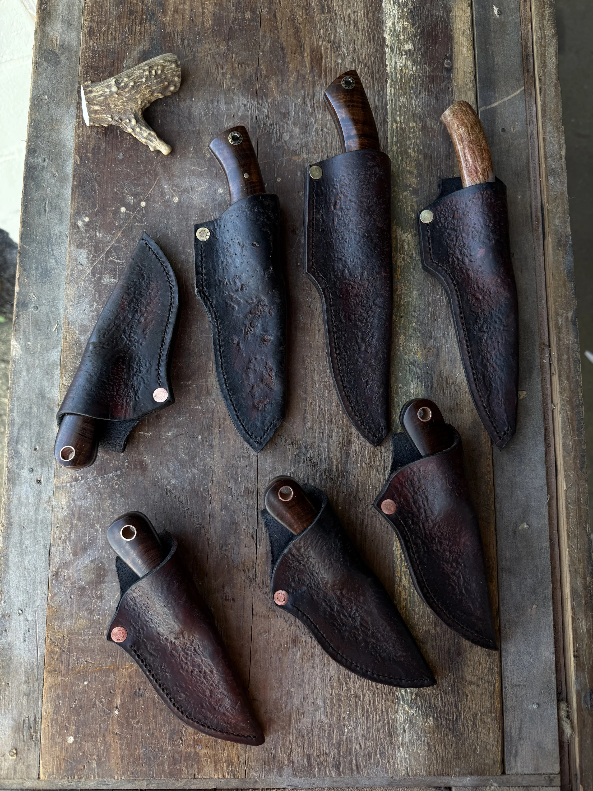 Six handmade knives with wooden handles sheathed in black leather covers displayed on a wooden surface, with a small piece of driftwood or coral in the top left corner.
