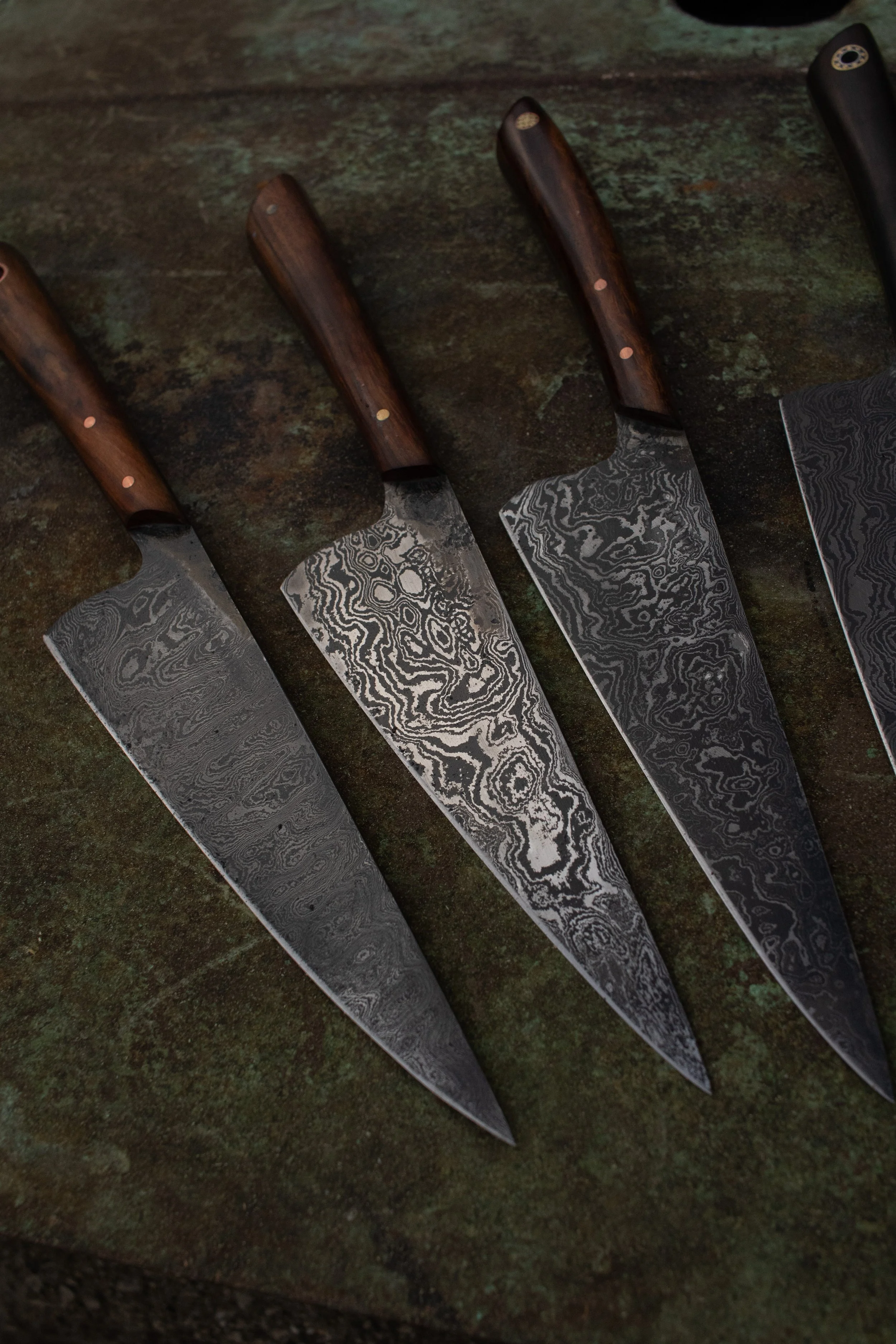 Four kitchen knives with wooden handles and patterned blades arranged diagonally on a rusted metal surface.