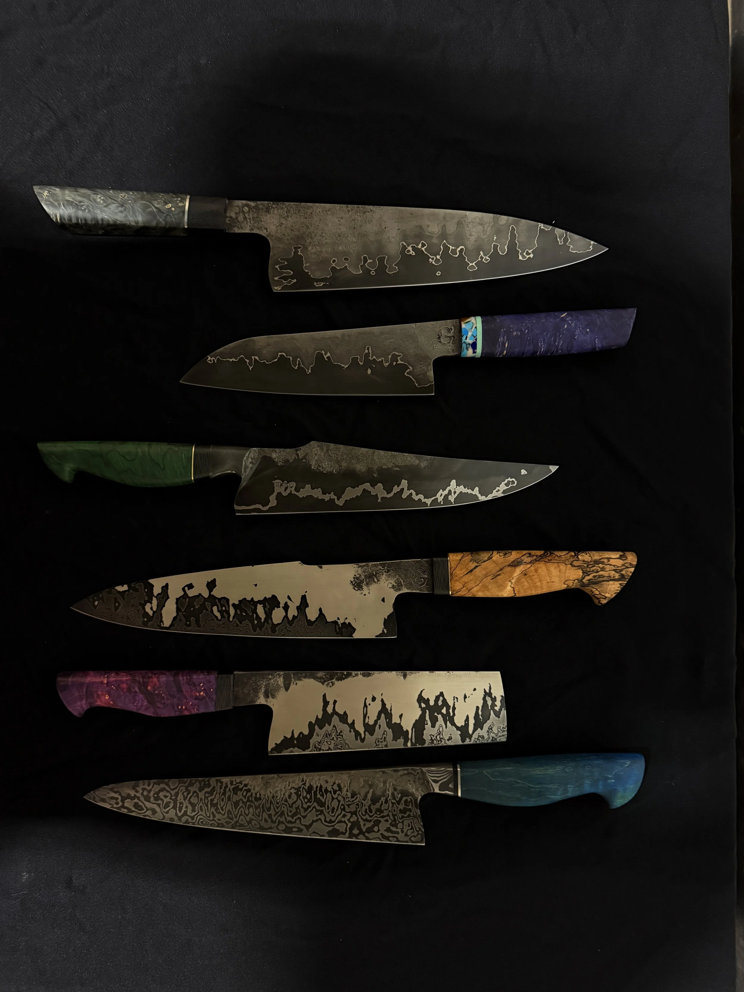 Six handcrafted custom kitchen knives with various patterned handles, arranged horizontally on a black surface.