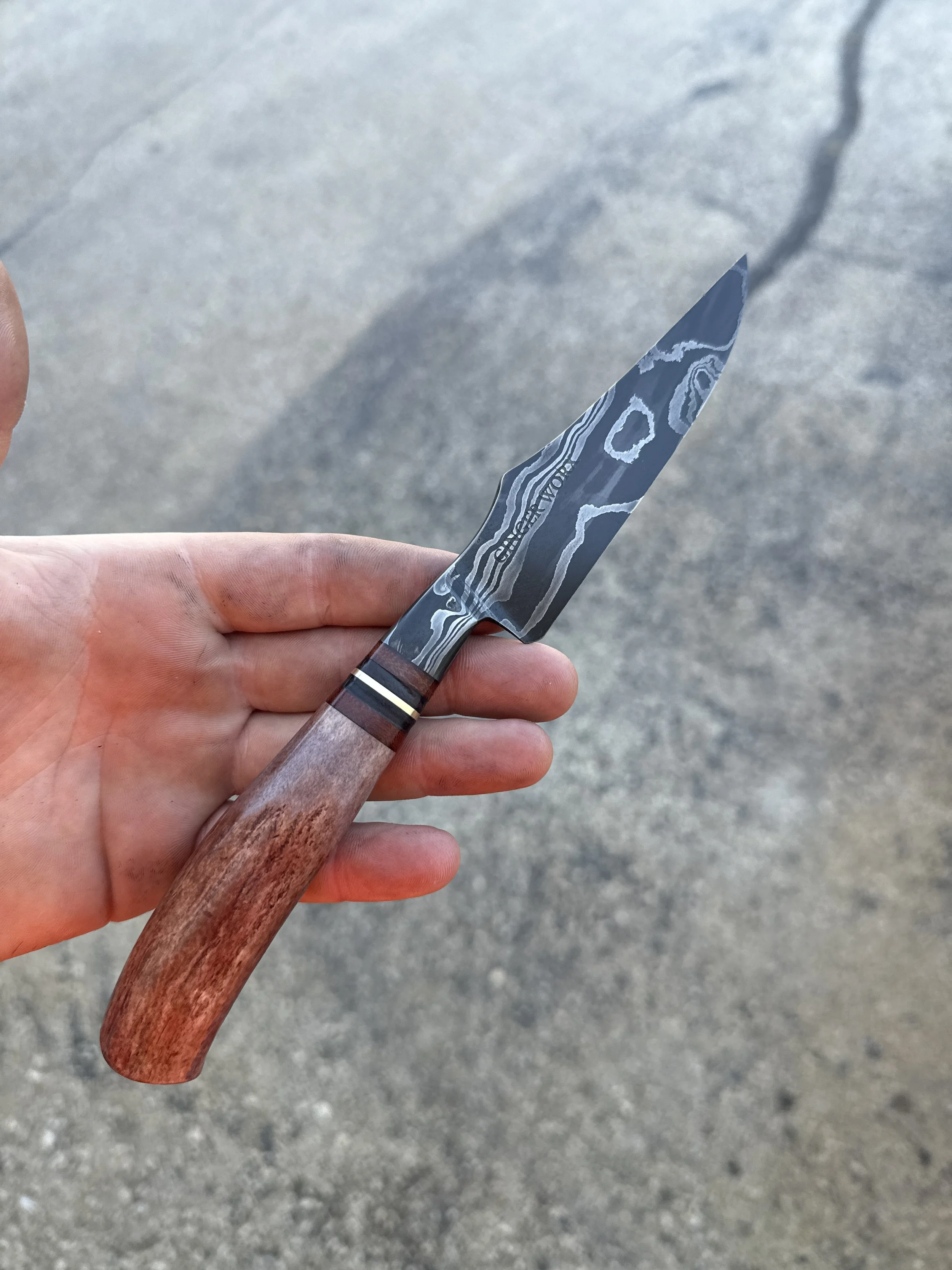 A hand holding a Damascus steel knife with a wooden handle over a gray concrete surface.