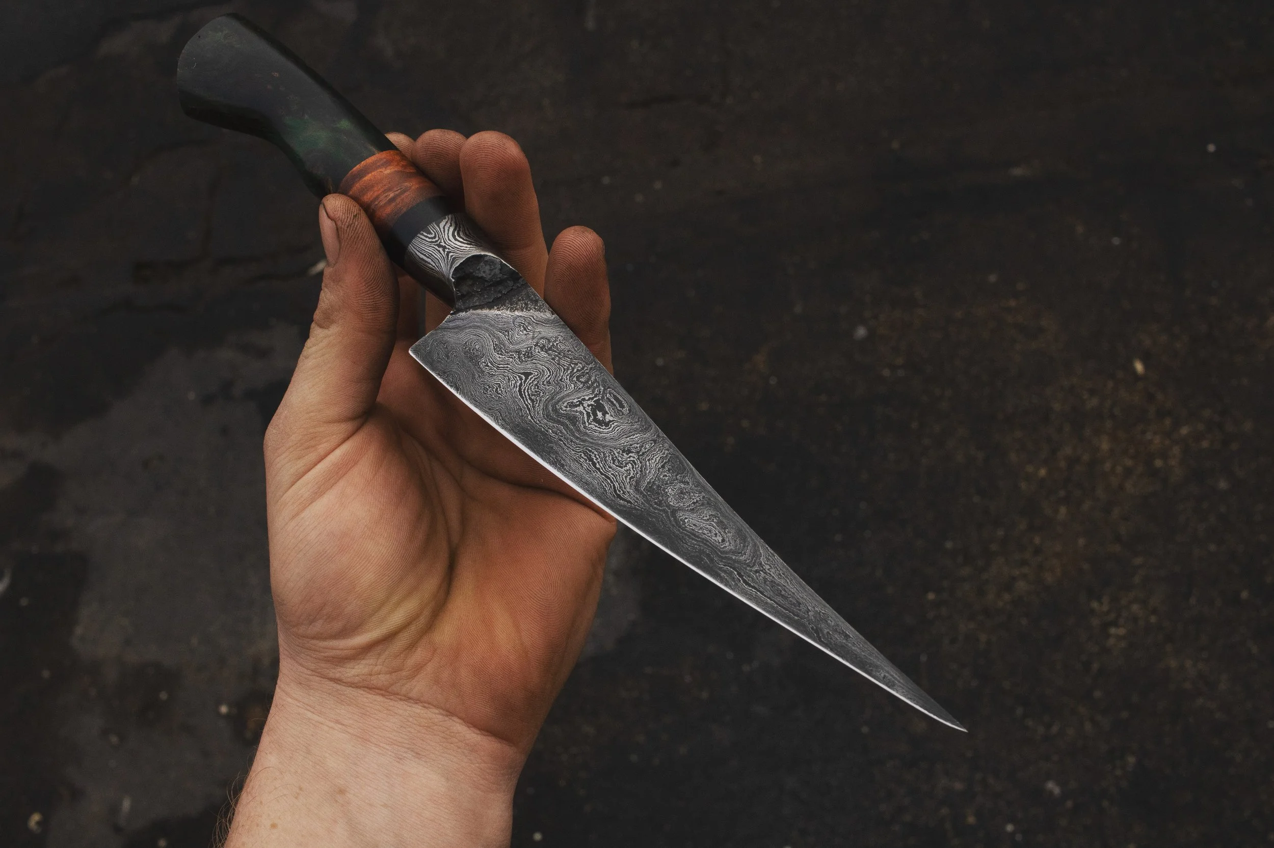Hand holding a Damascus steel kitchen knife with a wooden handle over a dark textured surface.