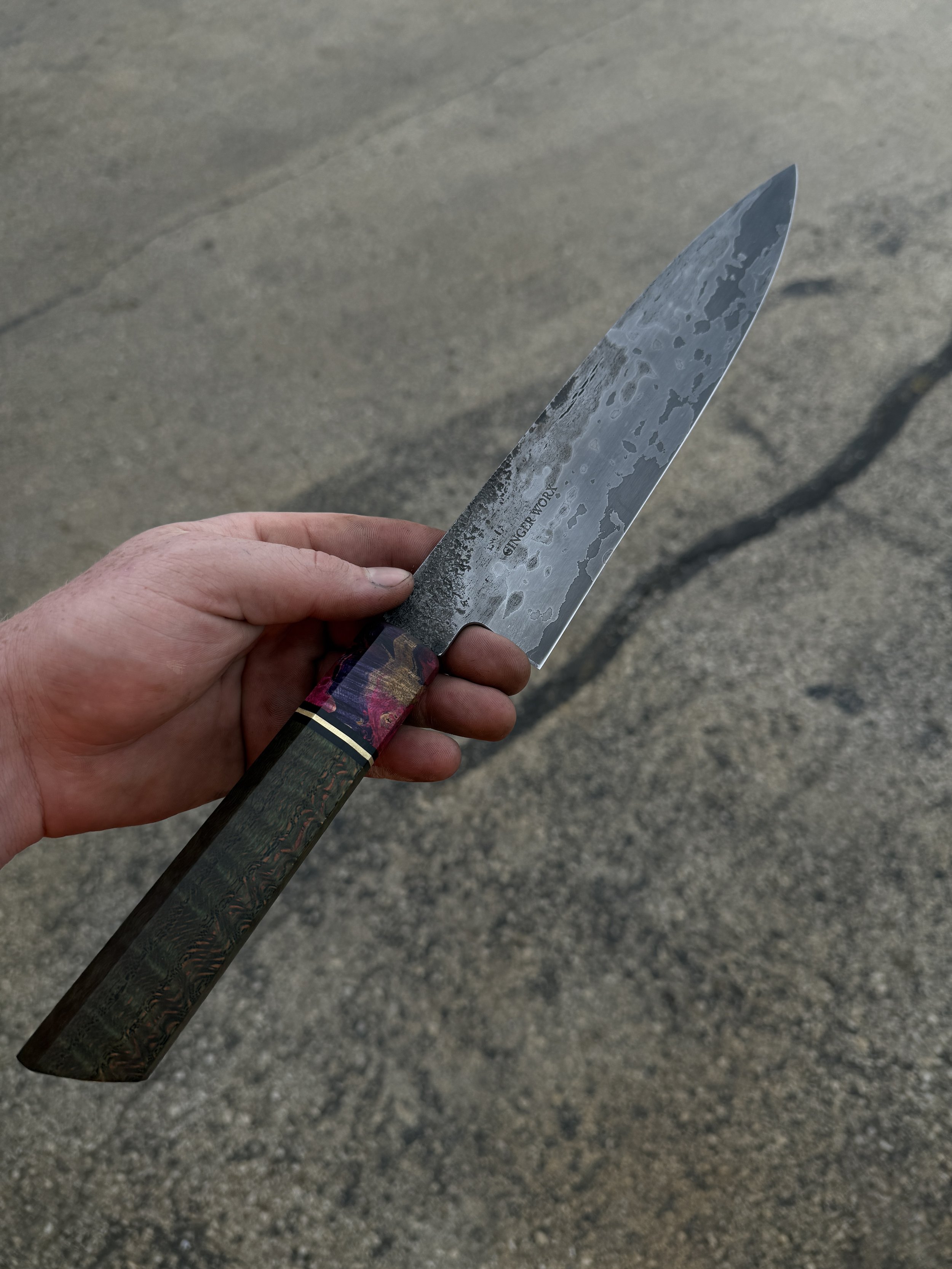 A hand holding a patterned chef's knife with a textured handle and a Damascus steel blade, outdoors on a paved surface.
