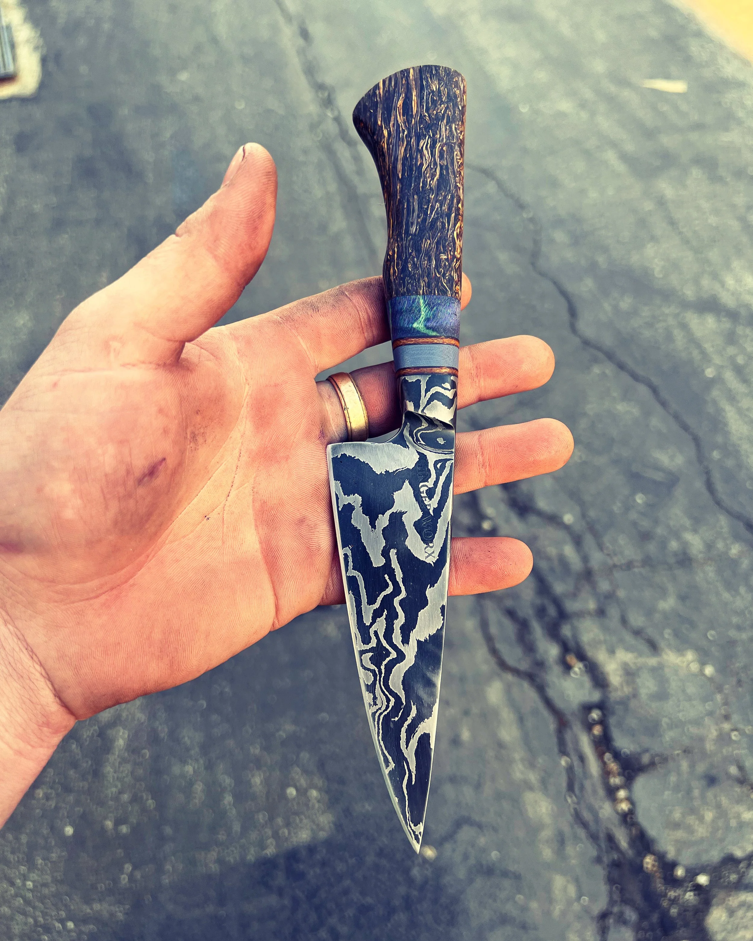A hand holding a Damascus steel knife with a textured, dark wooden handle, against a concrete background.