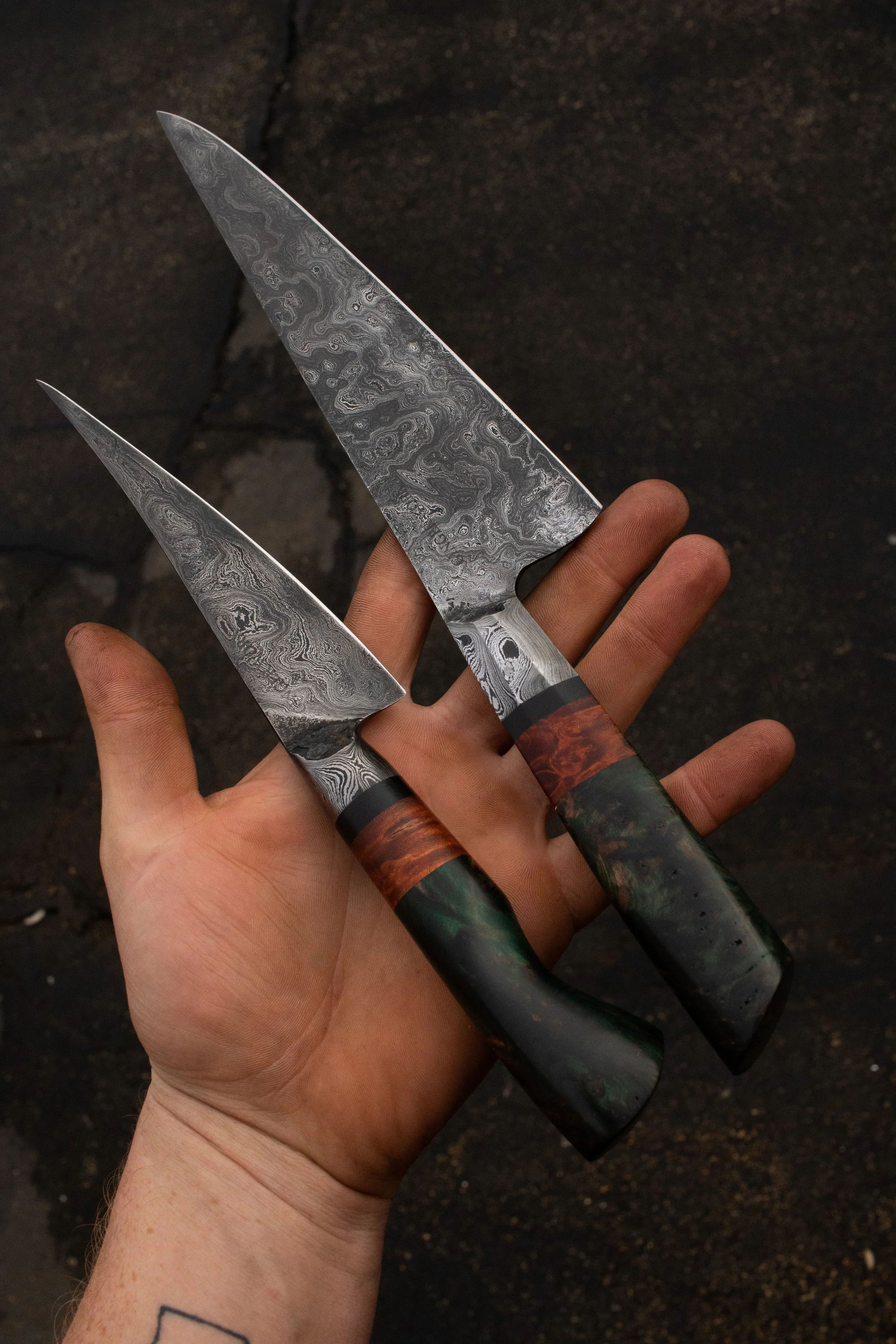 Two Damascus steel knives with dark multi-colored handles held over a dark ground.