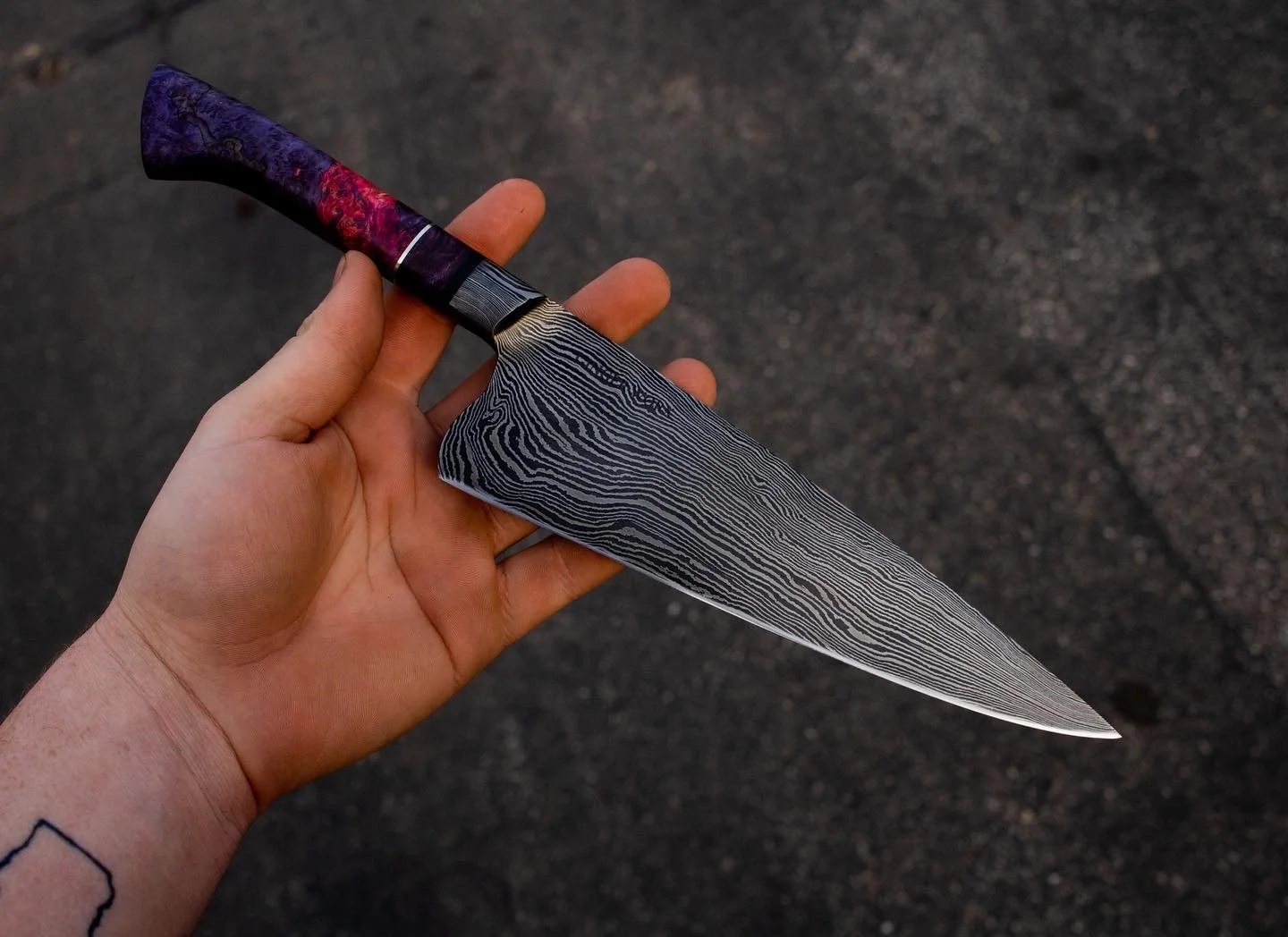 A hand holding a Damascus steel knife with a colorful handle, against a dark asphalt background.