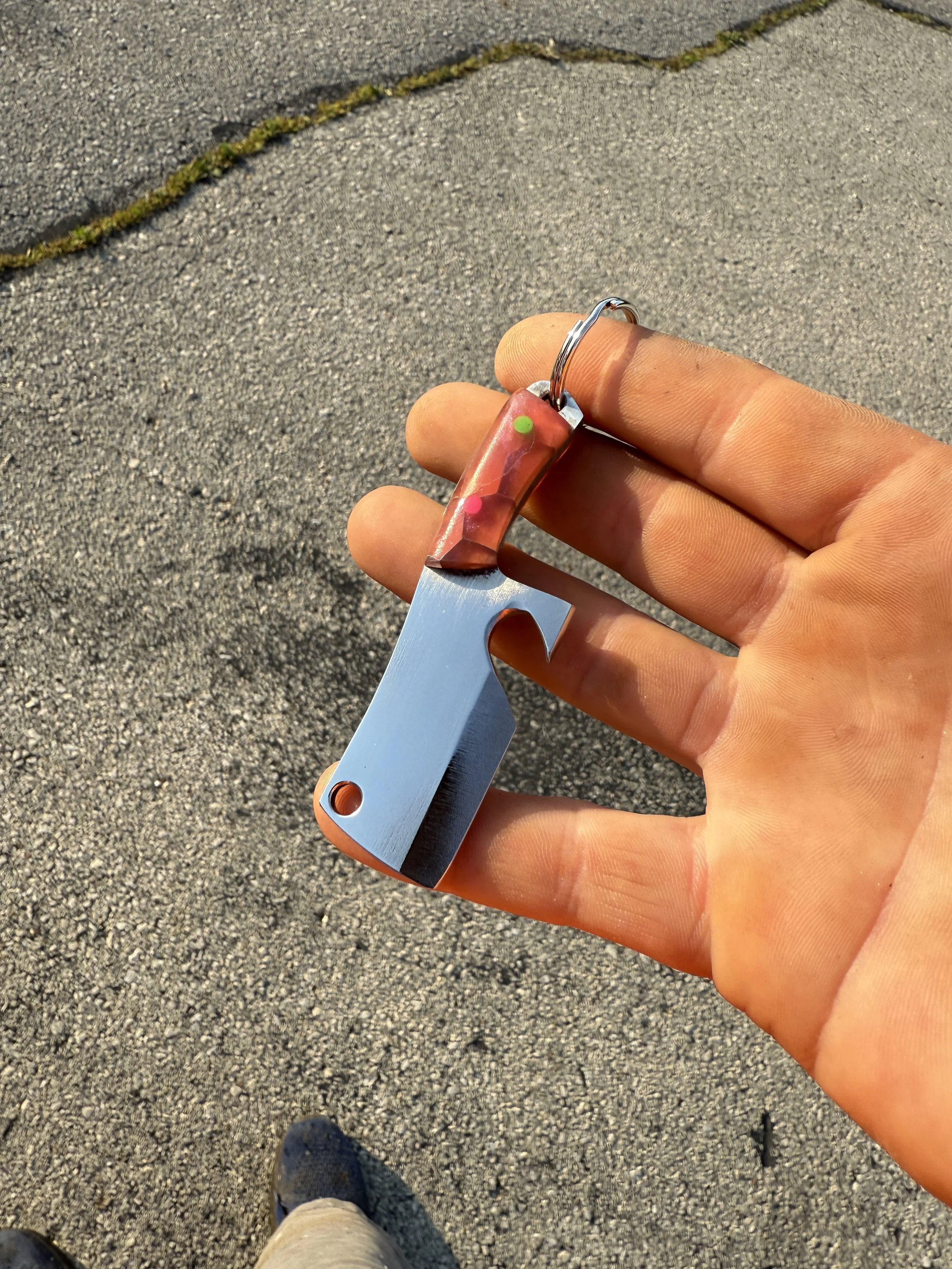 A hand holding a small handcrafted knife with a metal blade and a handle made of resin with embedded colorful dots, outdoors on a pavement.