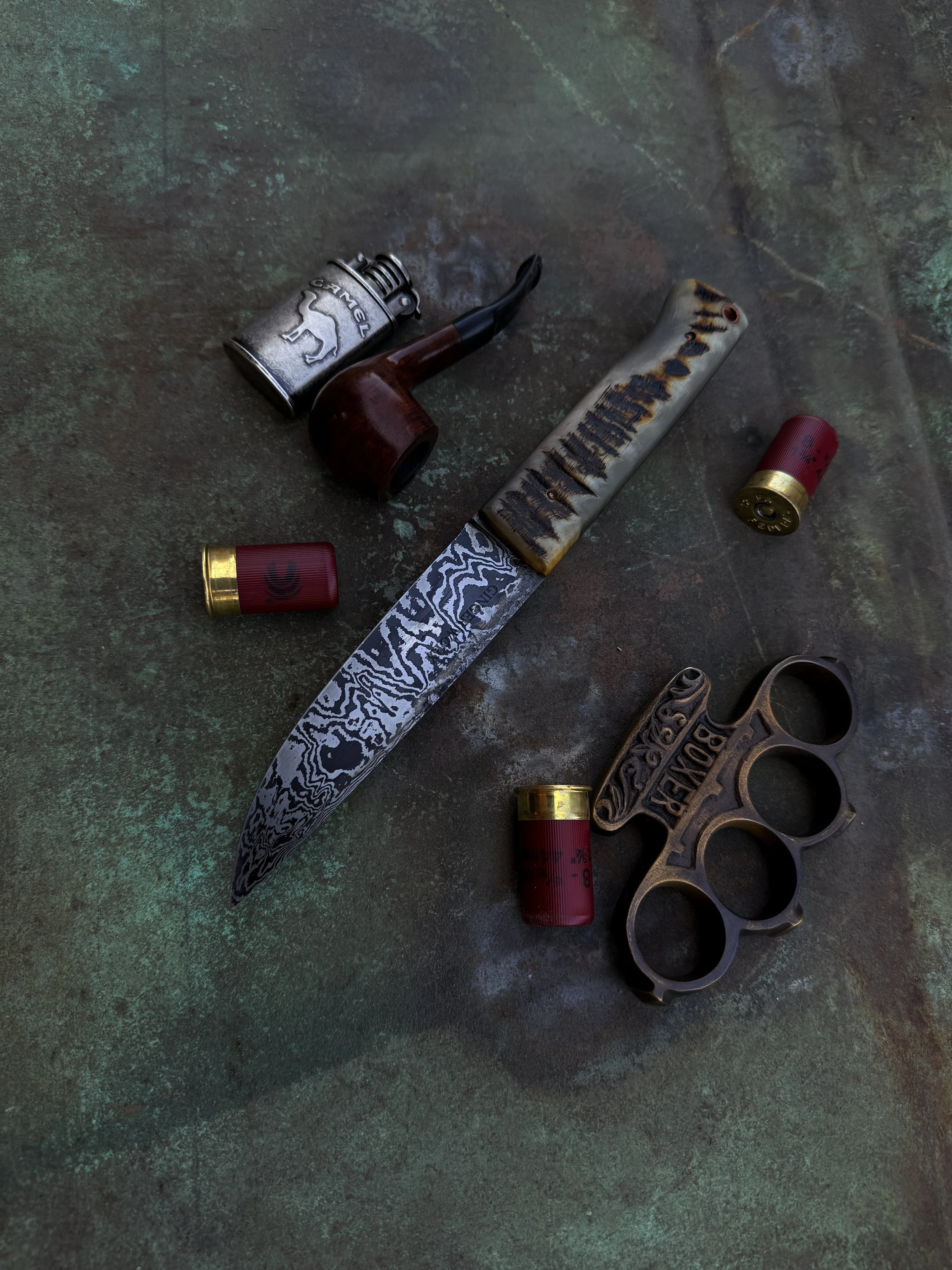 A collection of weapons and ammunition on a dark textured surface, including a Damascus steel knife with a bone handle, a brass knuckle-shaped brass tool, a smoking pipe, a small silver flask with a dog engraving, and three shotgun shells.