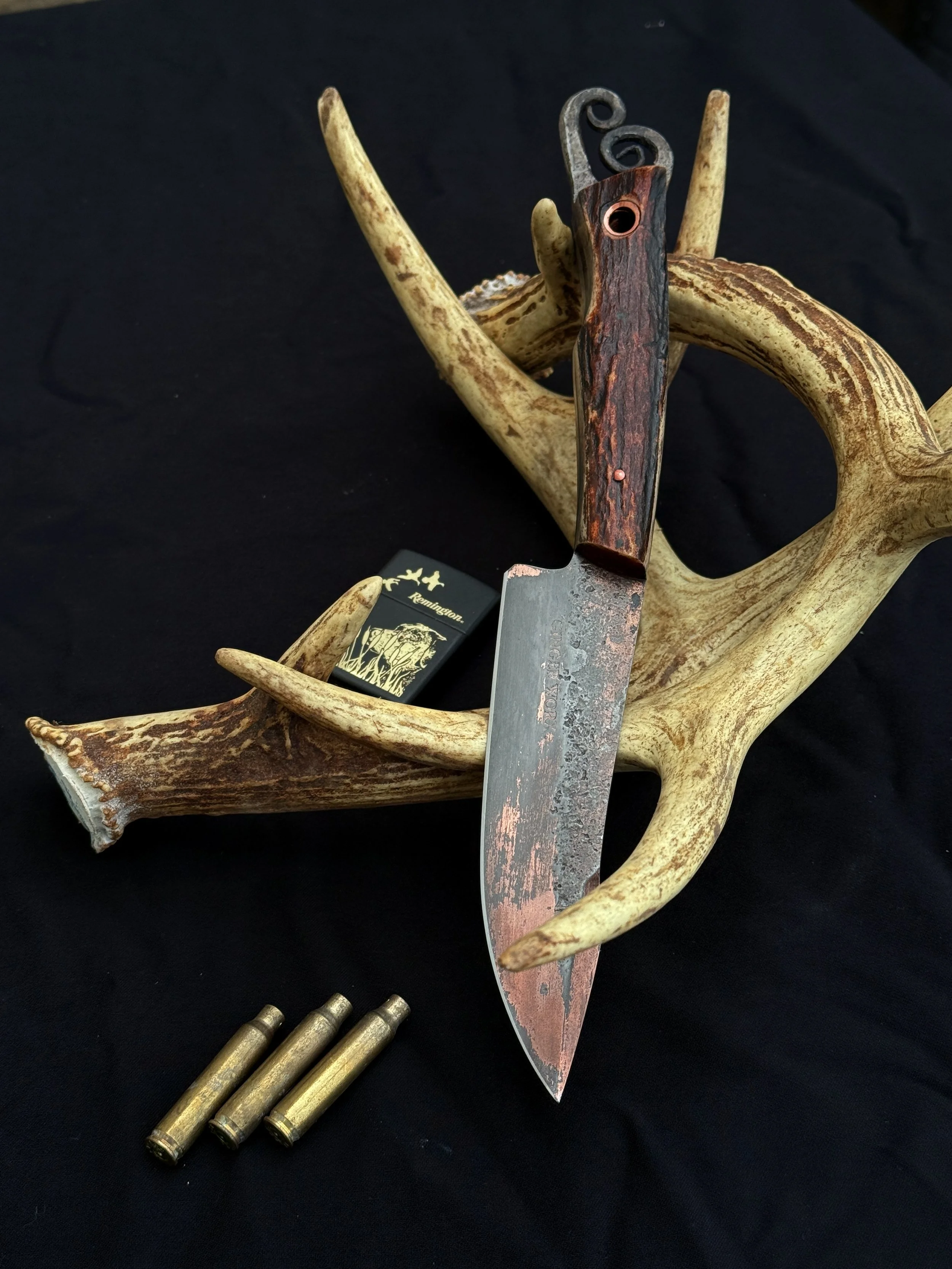 A rusty hunting knife with a wooden handle rests on an antler, with three spent bullet casings nearby, all placed on a black background.