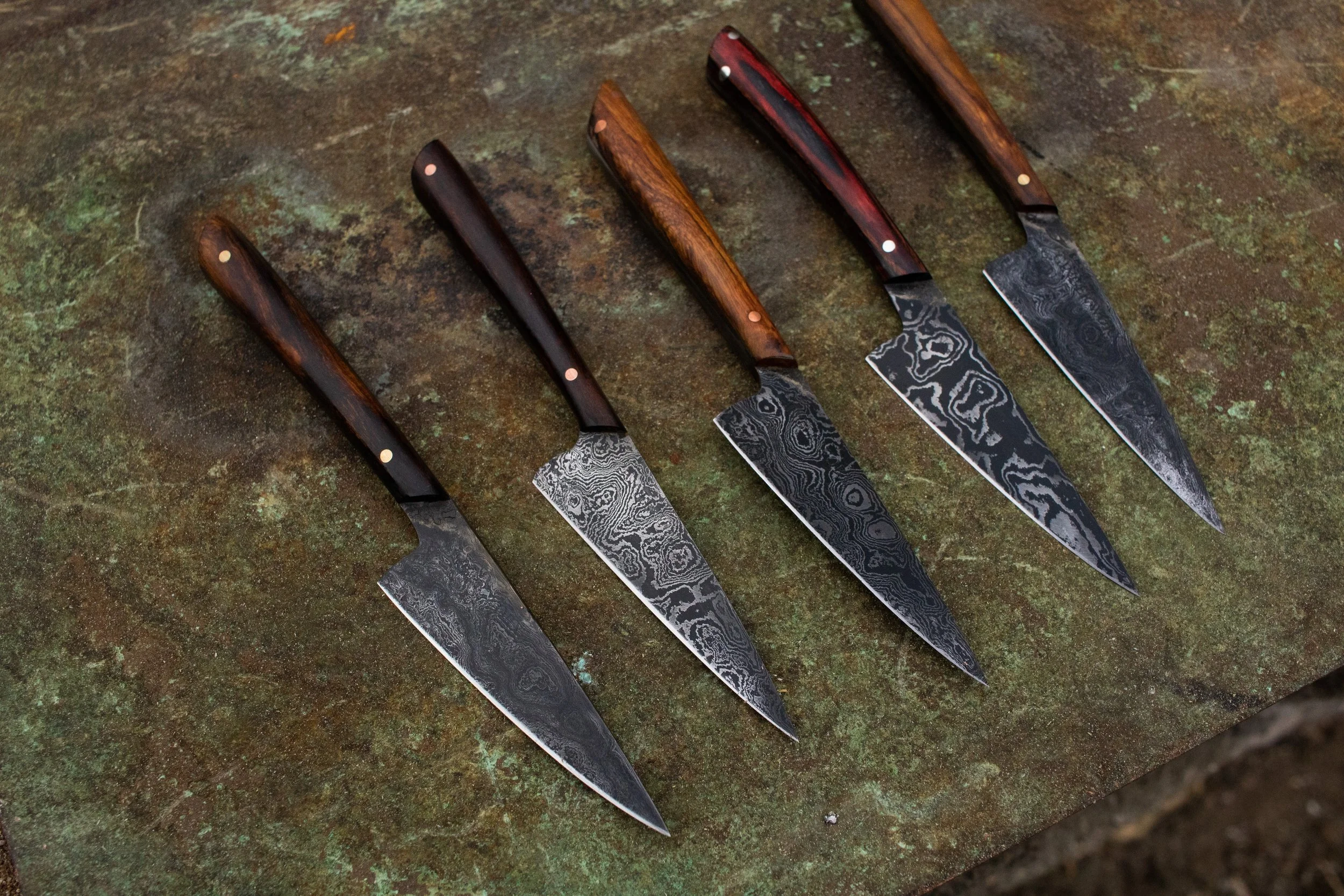 Five handcrafted knives with Damascus steel blades and wooden handles, arranged in a row on a rusty, textured surface.