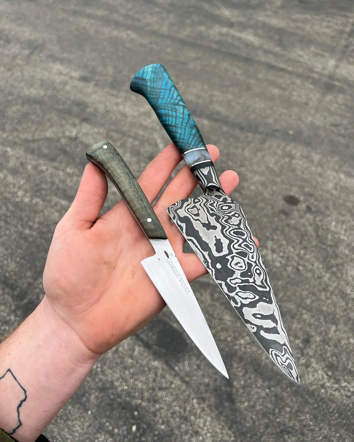 A hand holding two ornate knives with patterned blades and handles, with a plain asphalt surface in the background.