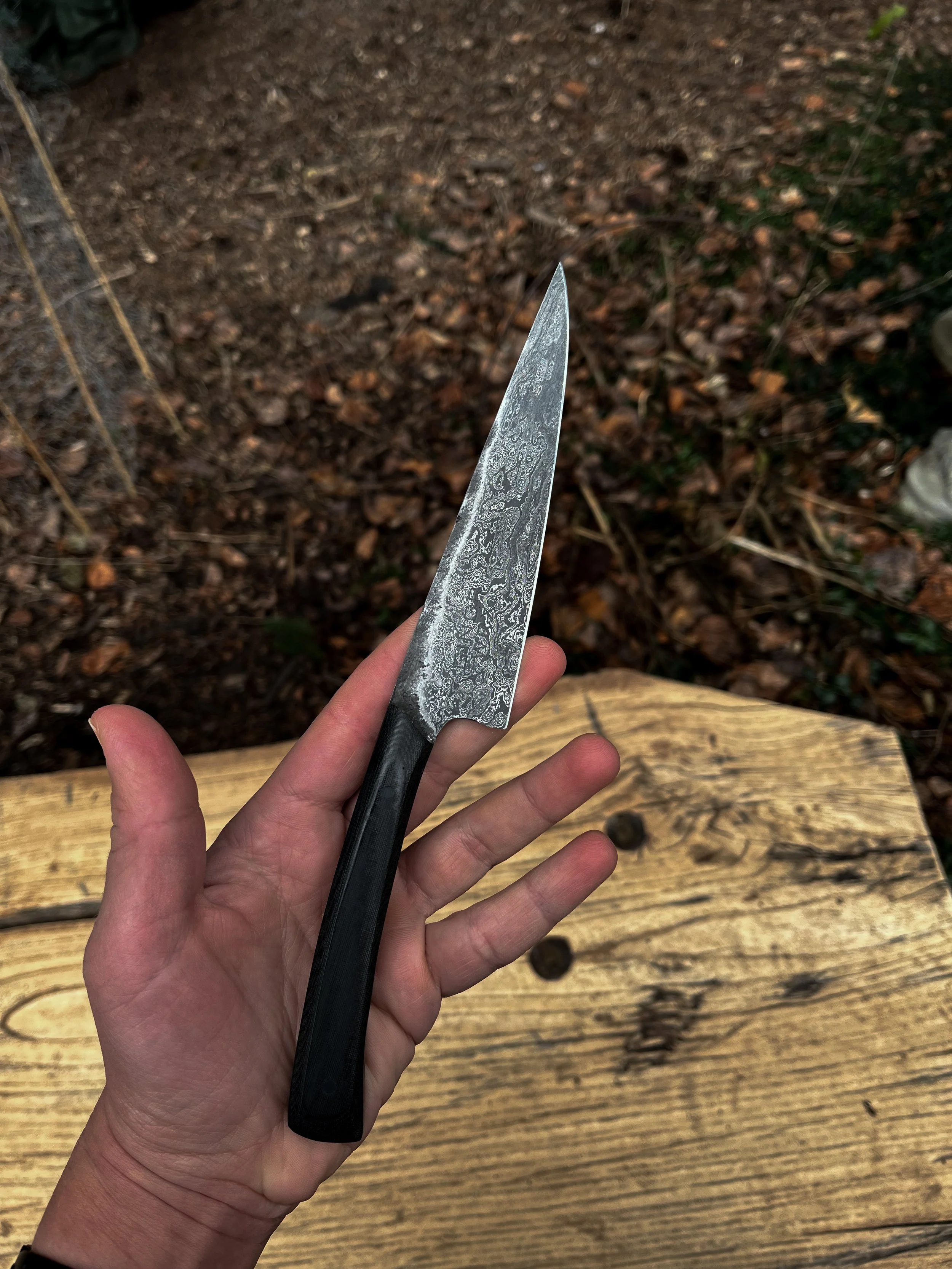 A person's hand holding a Damascus steel kitchen knife over a wooden surface outdoors, with soil and leaves in the background.