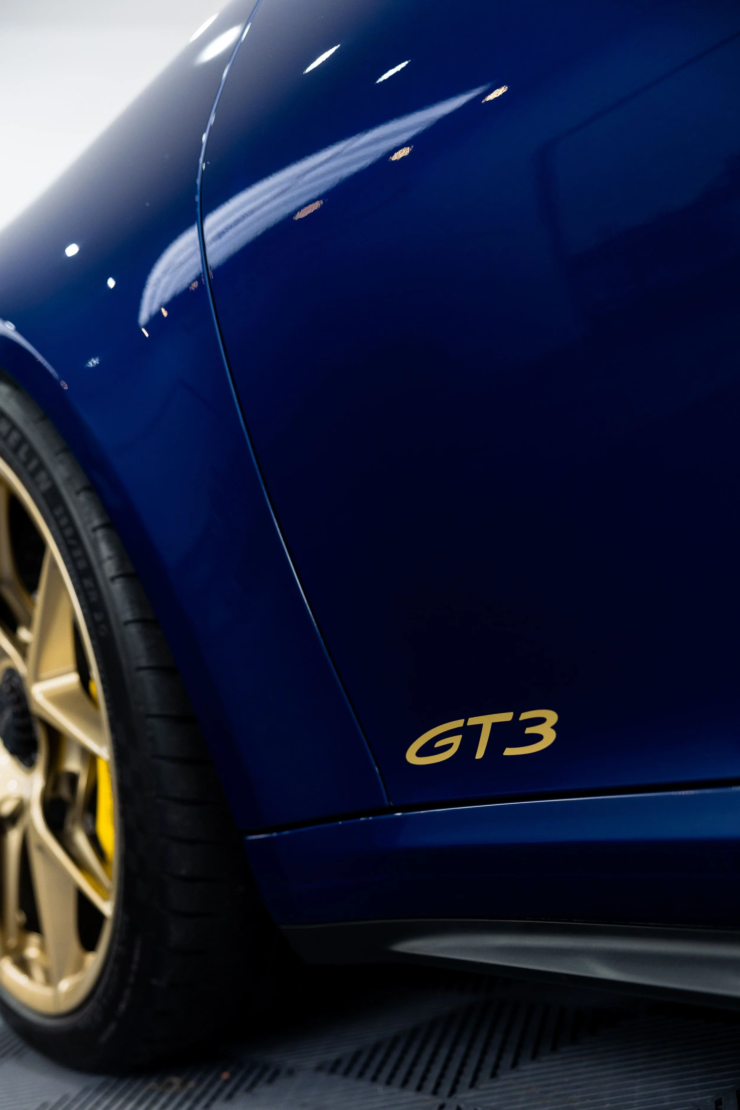 Close-up of blue Porsche 911 GT3 rear quarter panel with gold GT3 badge and gold wheel in detailing studio