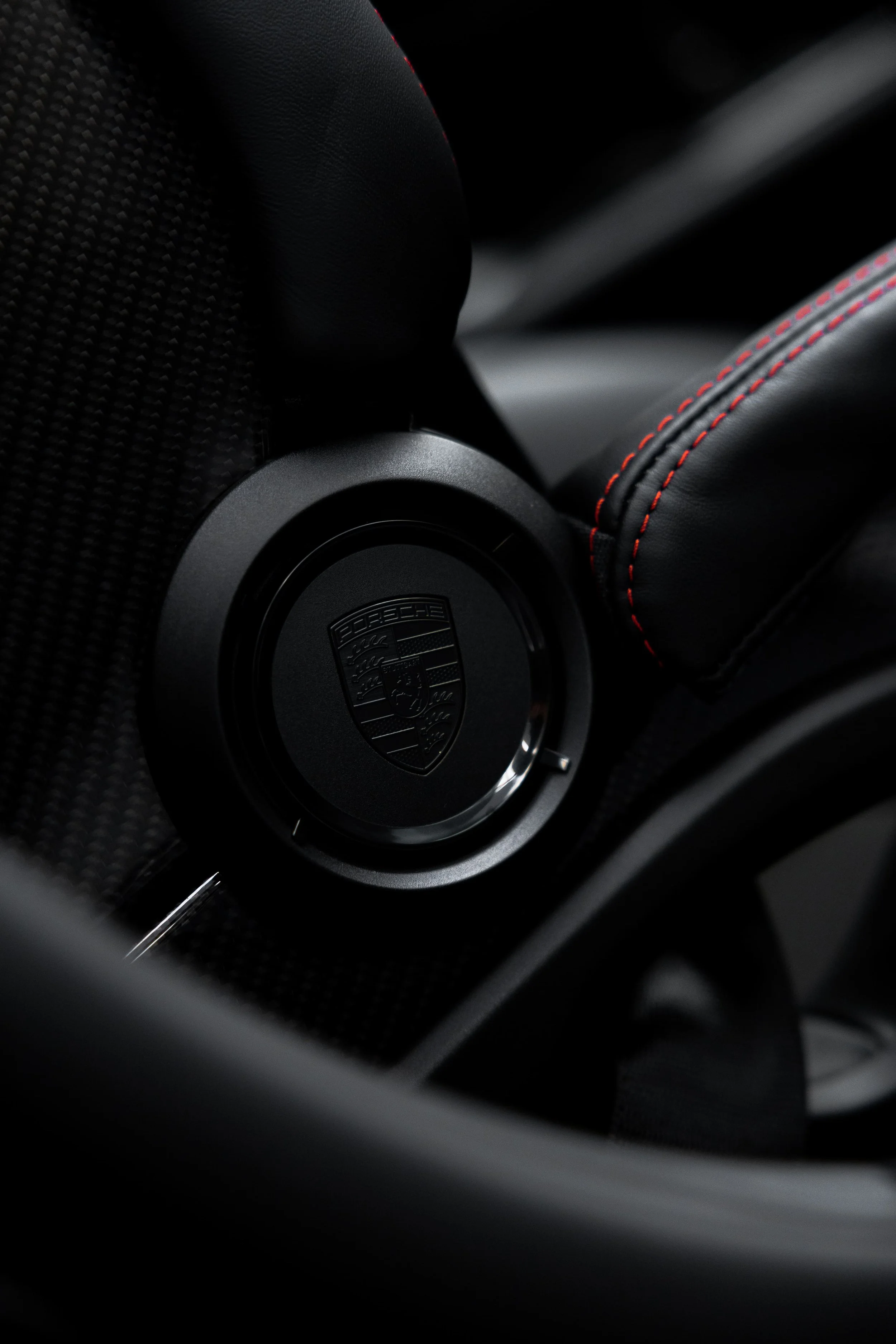 Close-up of Porsche 911 GT3 interior seat hinge with Porsche crest, carbon fibre finish, and red contrast stitching