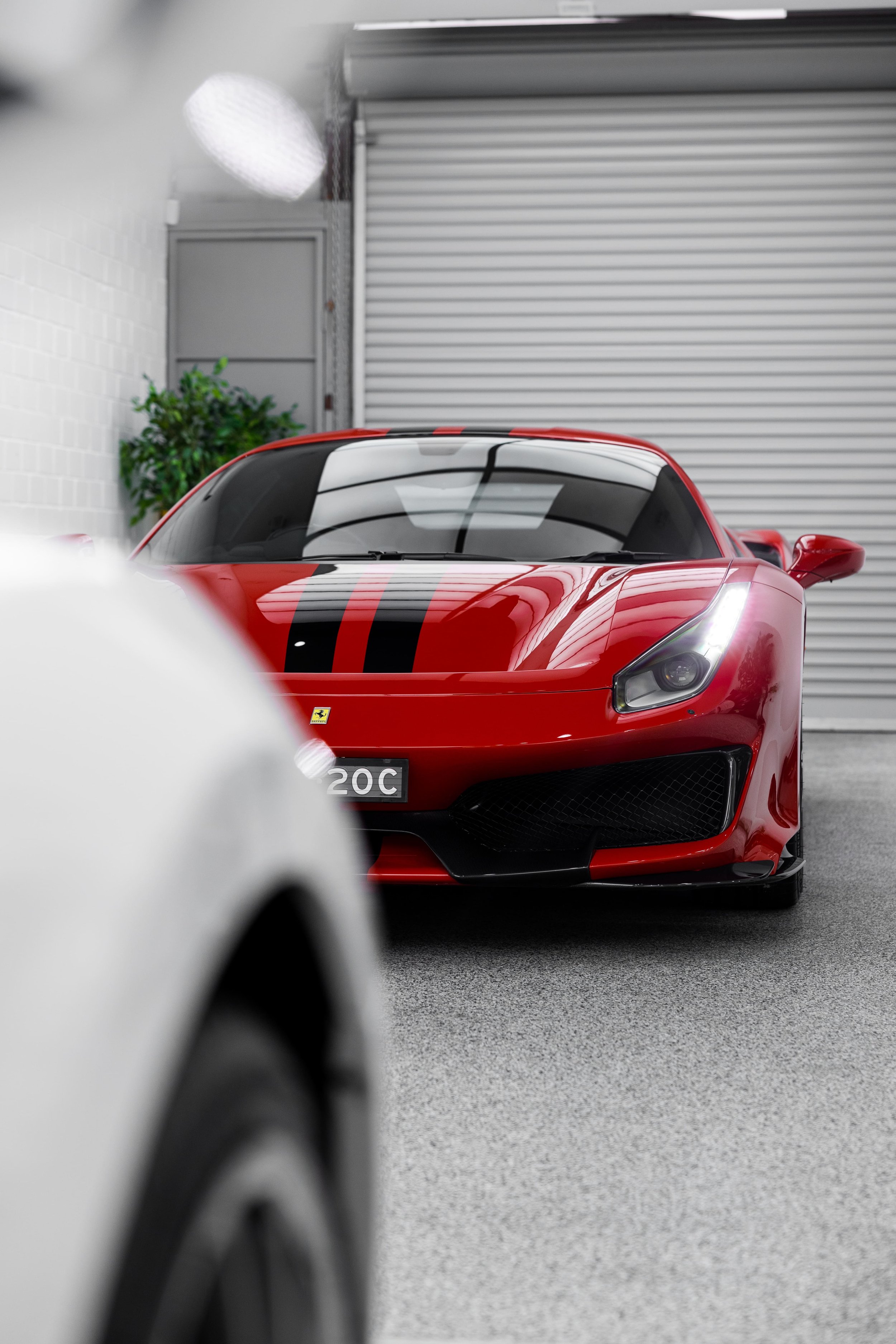 Front view of Ferrari 488 Pista with black racing stripes in detailing studio, partially framed by another vehicle in foreground