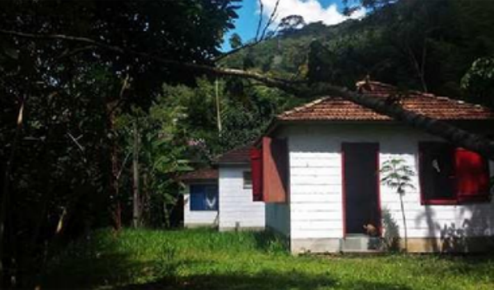 Small white house with red shutters and a tiled roof surrounded by greenery and trees, with a mountain in the background
