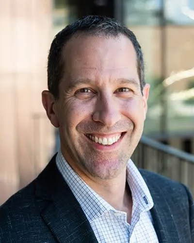 A smiling man in business attire standing outdoors with a blurred background of a building and railing.