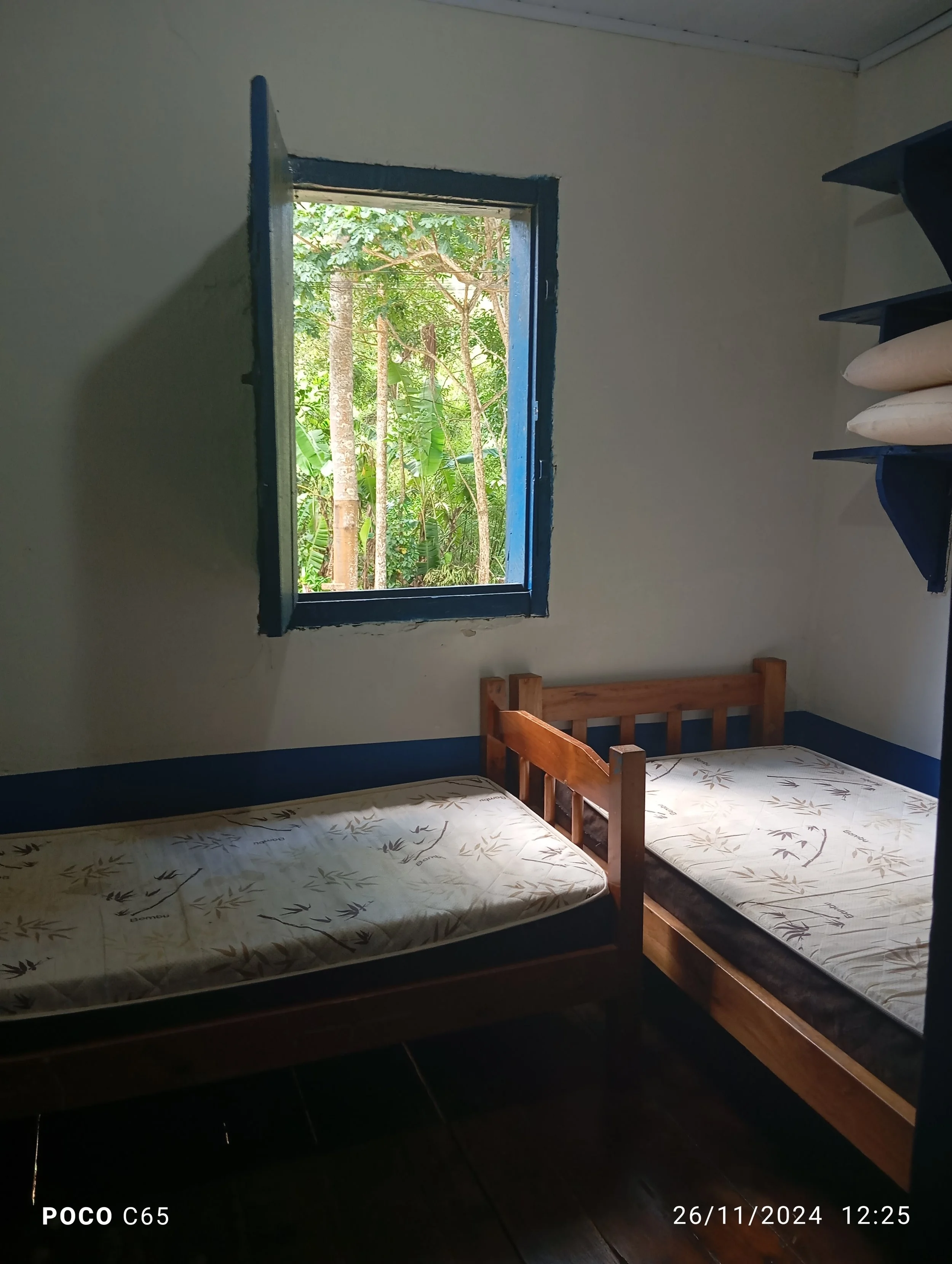 A small bedroom with two single beds, one with a headboard, a window showing green trees outside, and a shelf with pillows on the right.