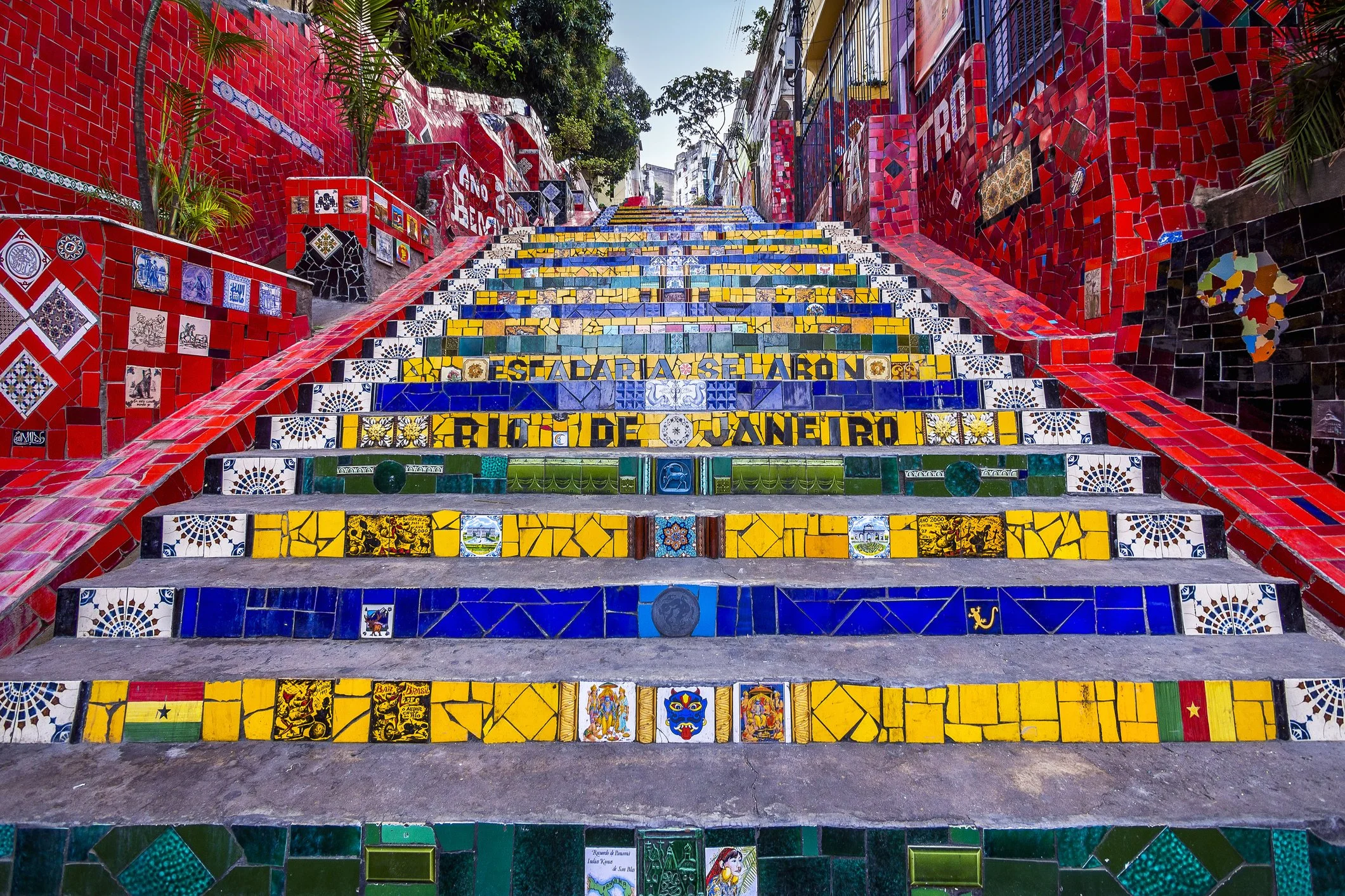Colorful mosaic stairs with a red wall on each side, decorated with various painted tiles and patterns, leading upward through a vibrant urban setting.