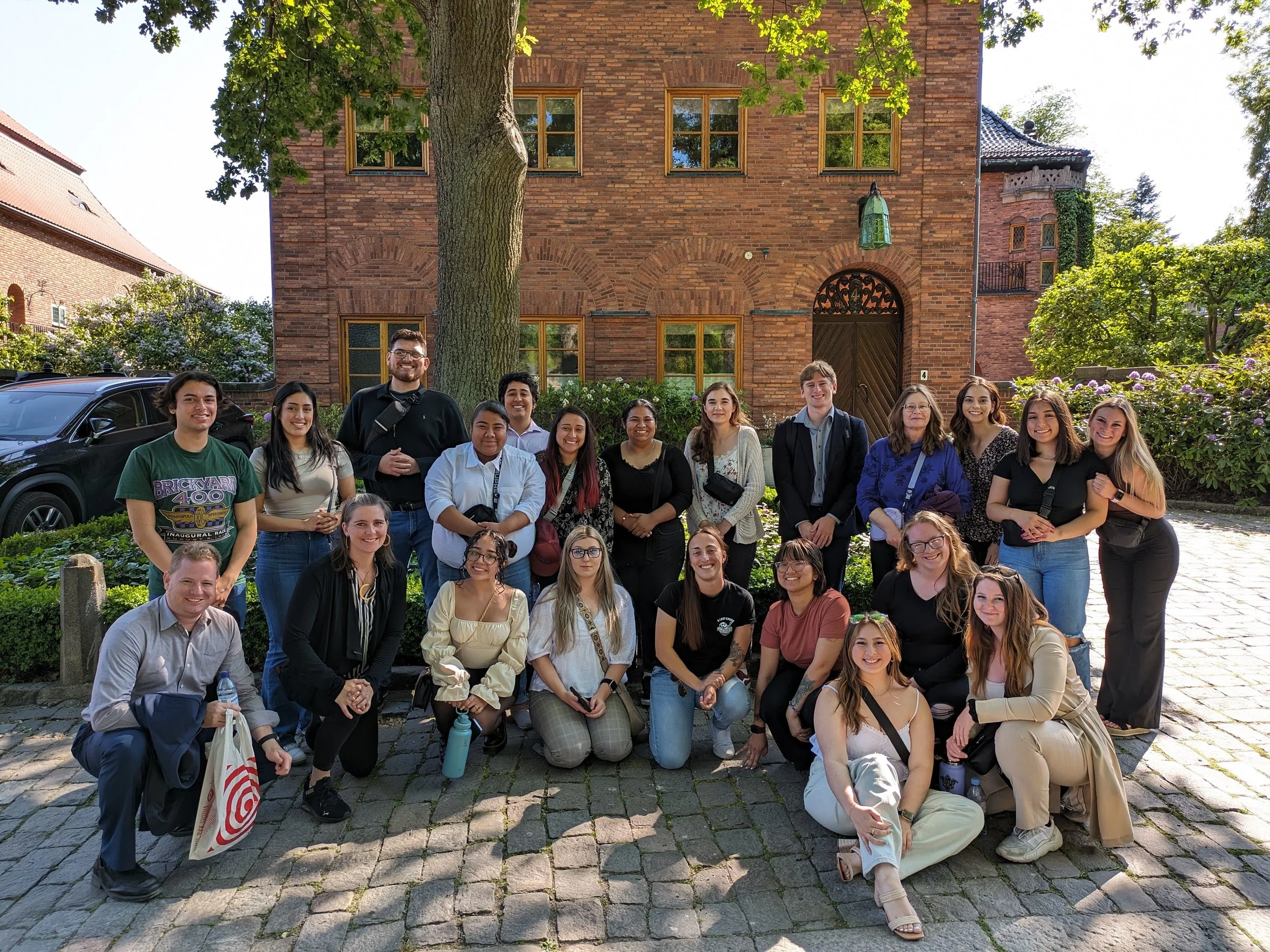 Group of young adults posing for a photo outdoors in front of a large brick building with a tree and greenery around.