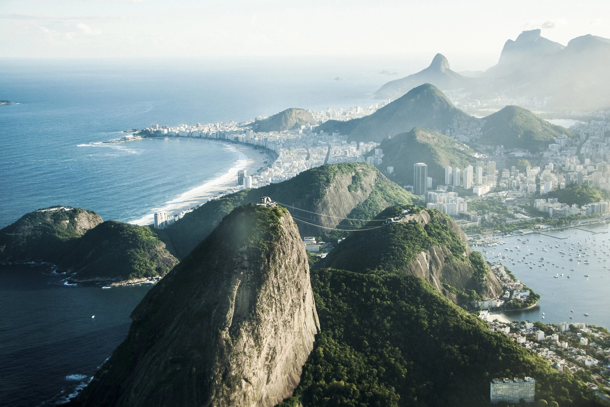 Aerial view of Rio de Janeiro with Sugarloaf Mountain and coastline, showing city buildings, beaches, and a harbor with boats.