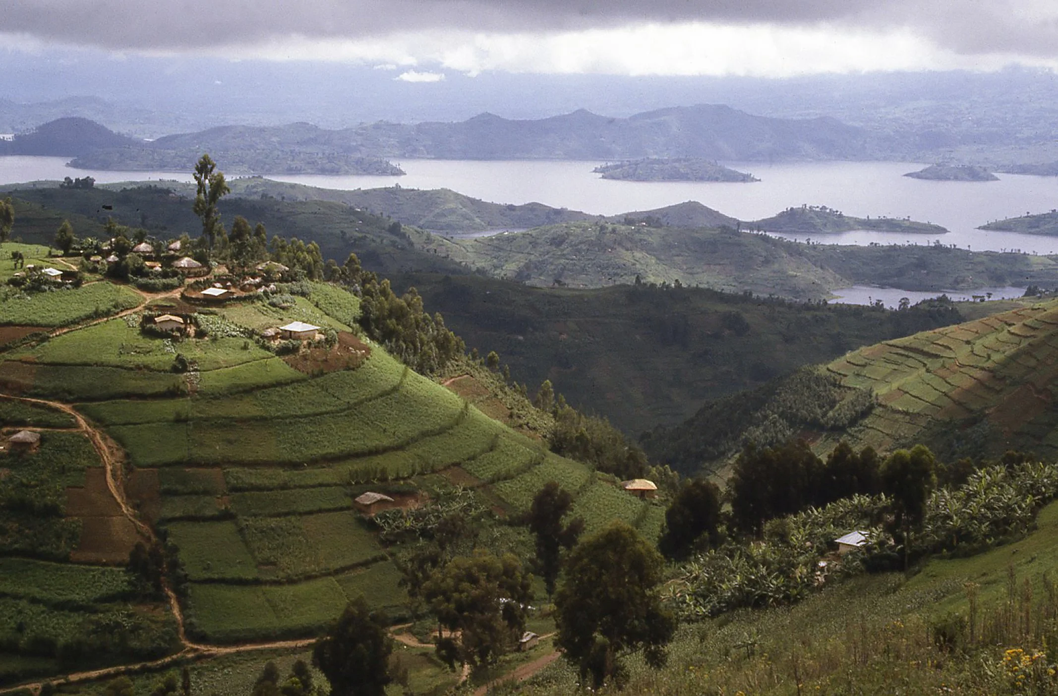 Lush green terraced hills with small houses overlooking a large lake and mountain ranges in the distance under cloudy sky.