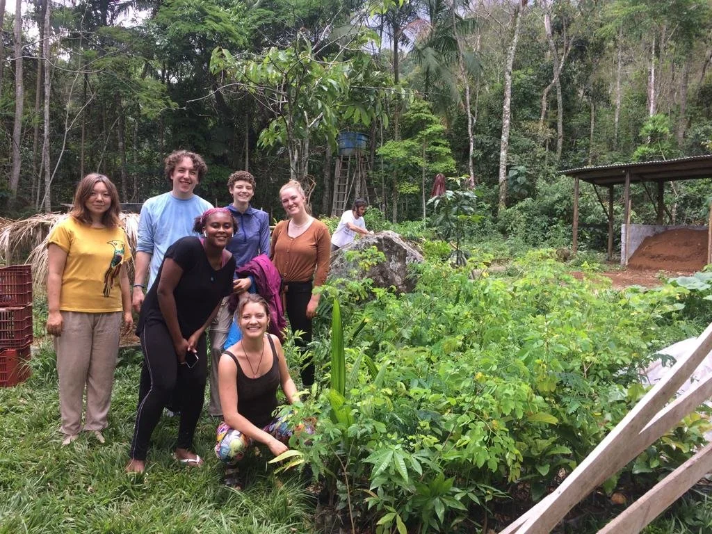 Group of seven smiling people standing and kneeling in a lush green garden with trees and plants, some structures and a person working in the background.