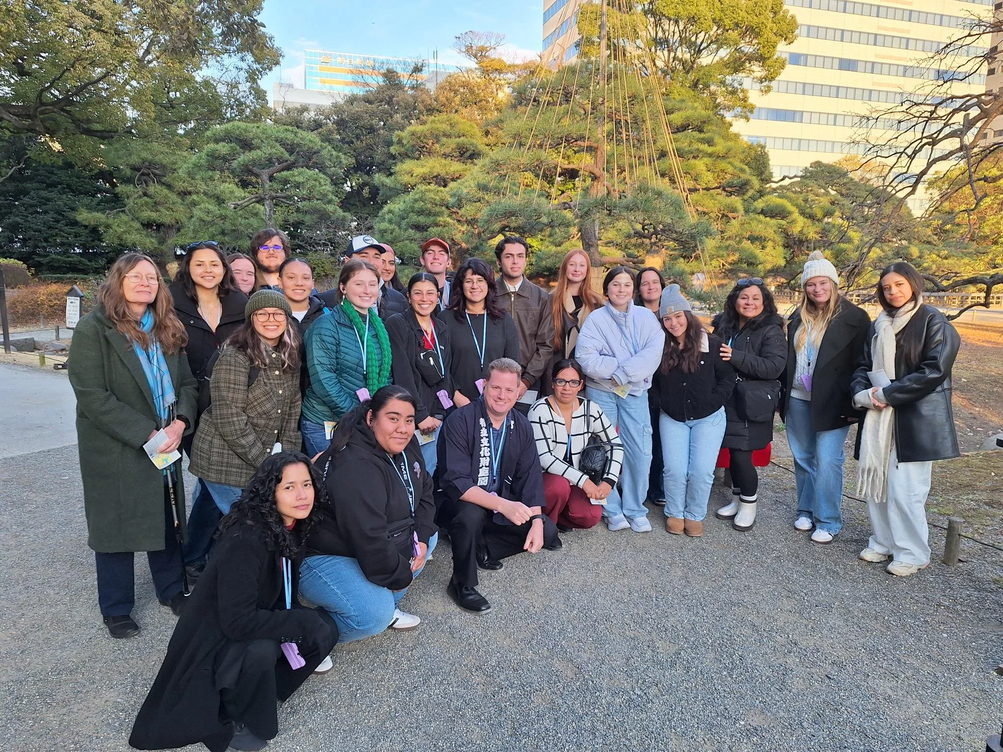 Group of people posing outdoors in front of a traditional Japanese garden with trees and a wooden structure with string supports.