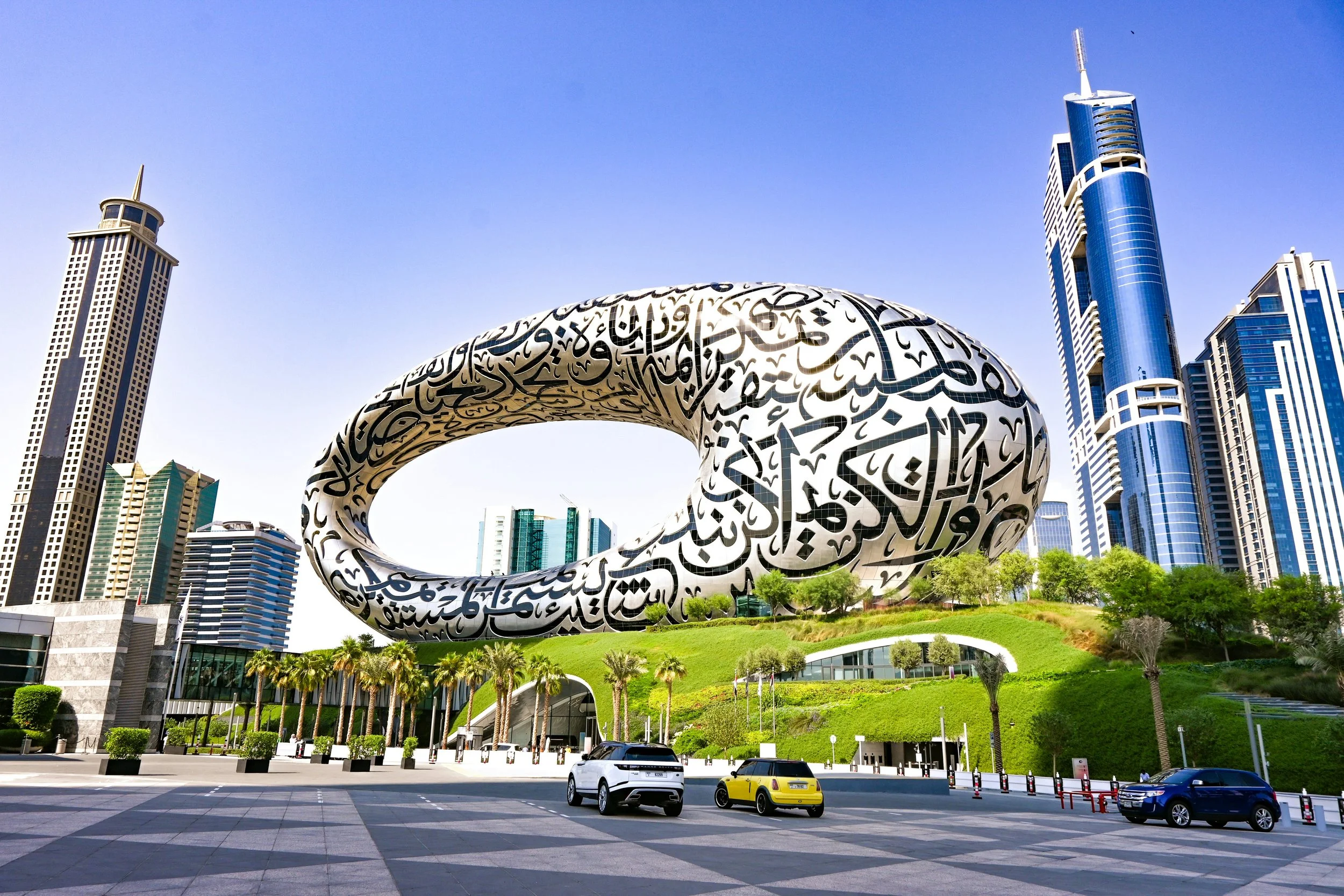Modern cityscape with a futuristic building featuring Arabic calligraphy, surrounded by tall skyscrapers, palm trees, and parked cars.