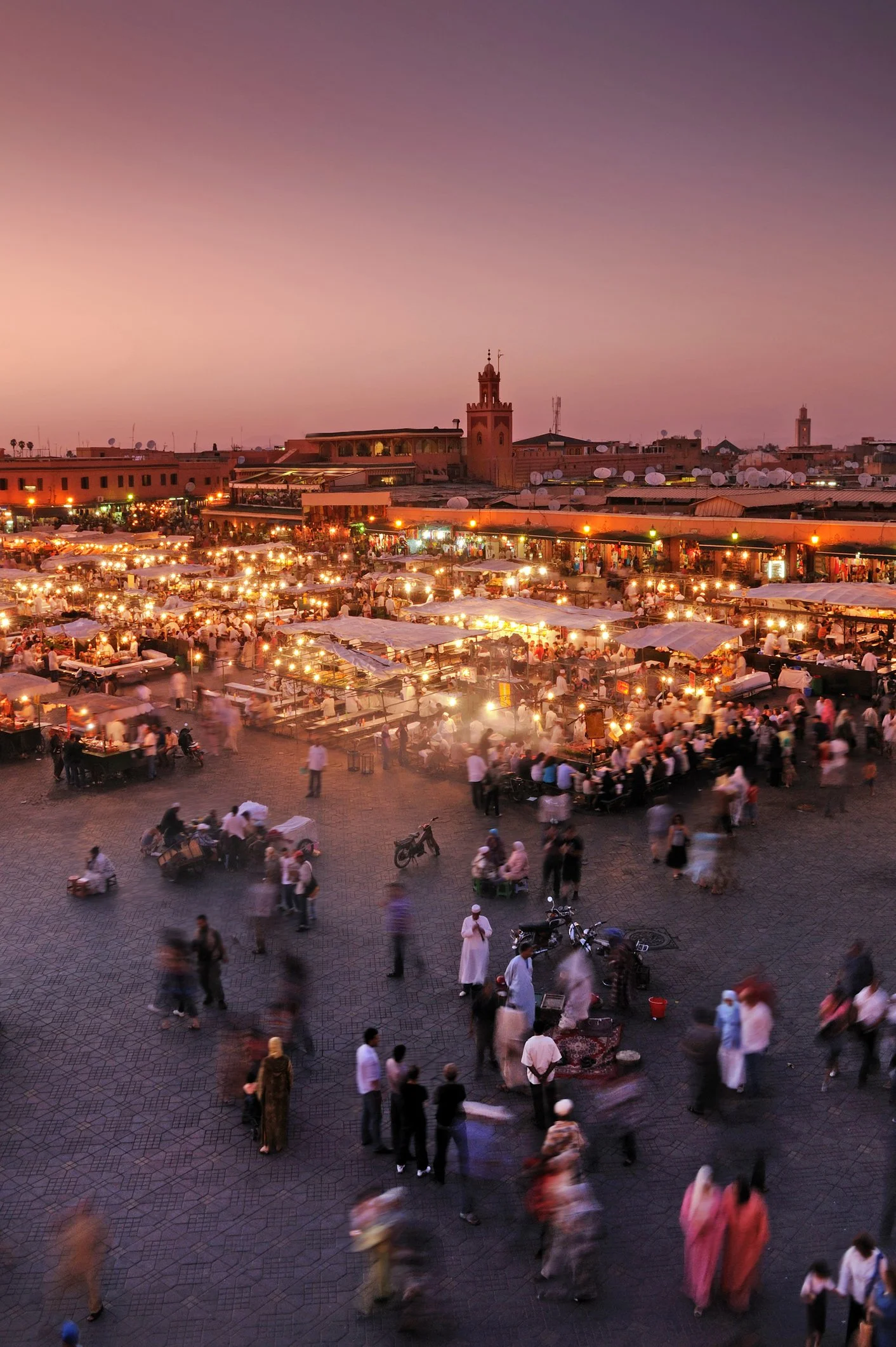 A bustling outdoor market at dusk, illuminated by string lights, with numerous vendors and people walking around, and a tall clock tower in the background beneath a purple sky.
