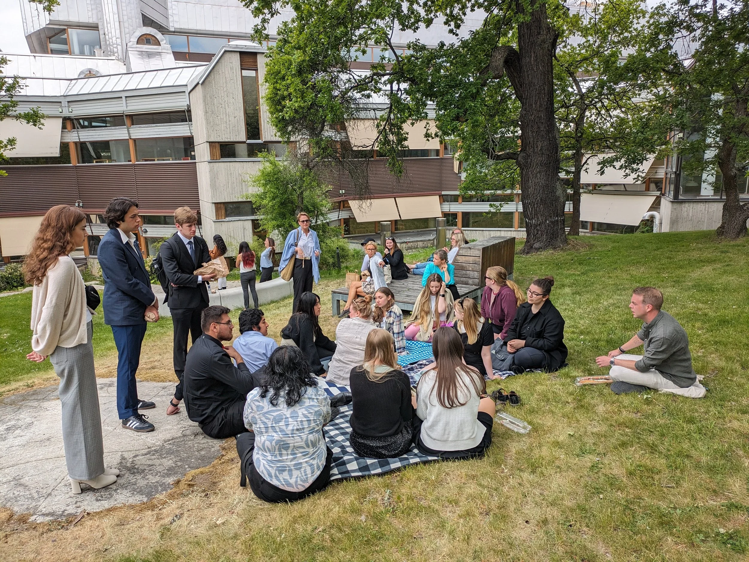 A group of people gathered outdoors on a grassy area, sitting on a checkered blanket and standing, participating in a discussion or event with trees and a modern building in the background.