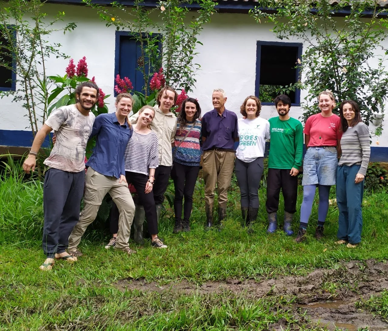 Group of eleven people standing outdoors on grass in front of a white house with blue trim, surrounded by green plants and pink flowers, smiling and posing for a photo.