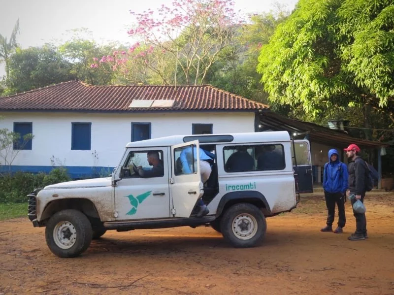A white off-road vehicle with people getting in and out standing on a dirt path near a white house with a red-tiled roof. Two people in jackets and hats stand nearby.