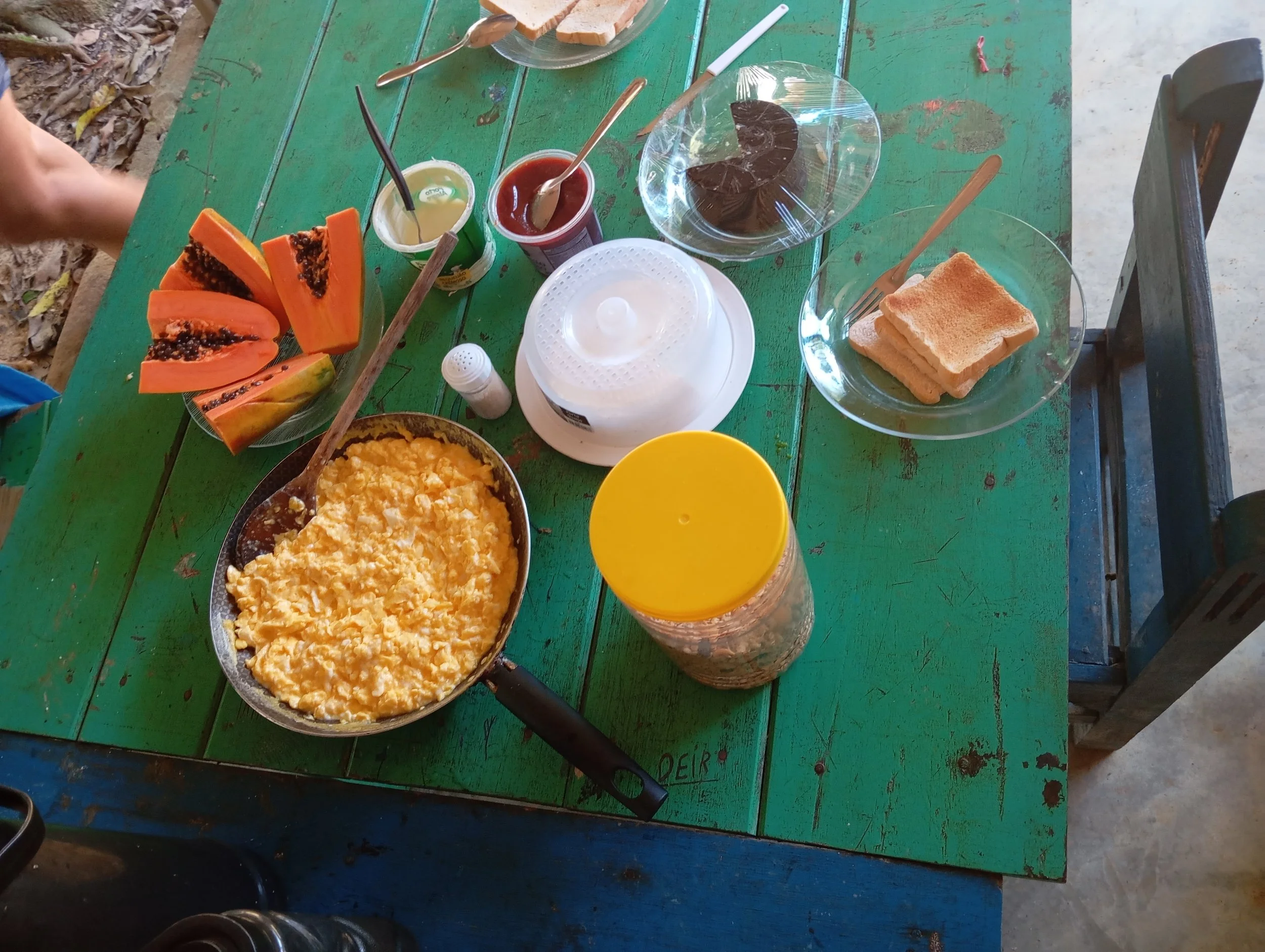 A green wooden table with breakfast items including scrambled eggs in a pan, sliced papaya on a plate, two bowls with bread slices and utensils, two jars of jam, a shaker, a white container, and a yellow cup with a beverage.