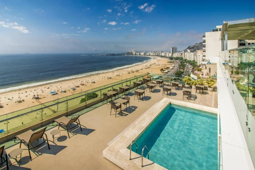 An oceanfront balcony with tables and chairs, overlooking a sandy beach and cityscape with high-rise buildings.