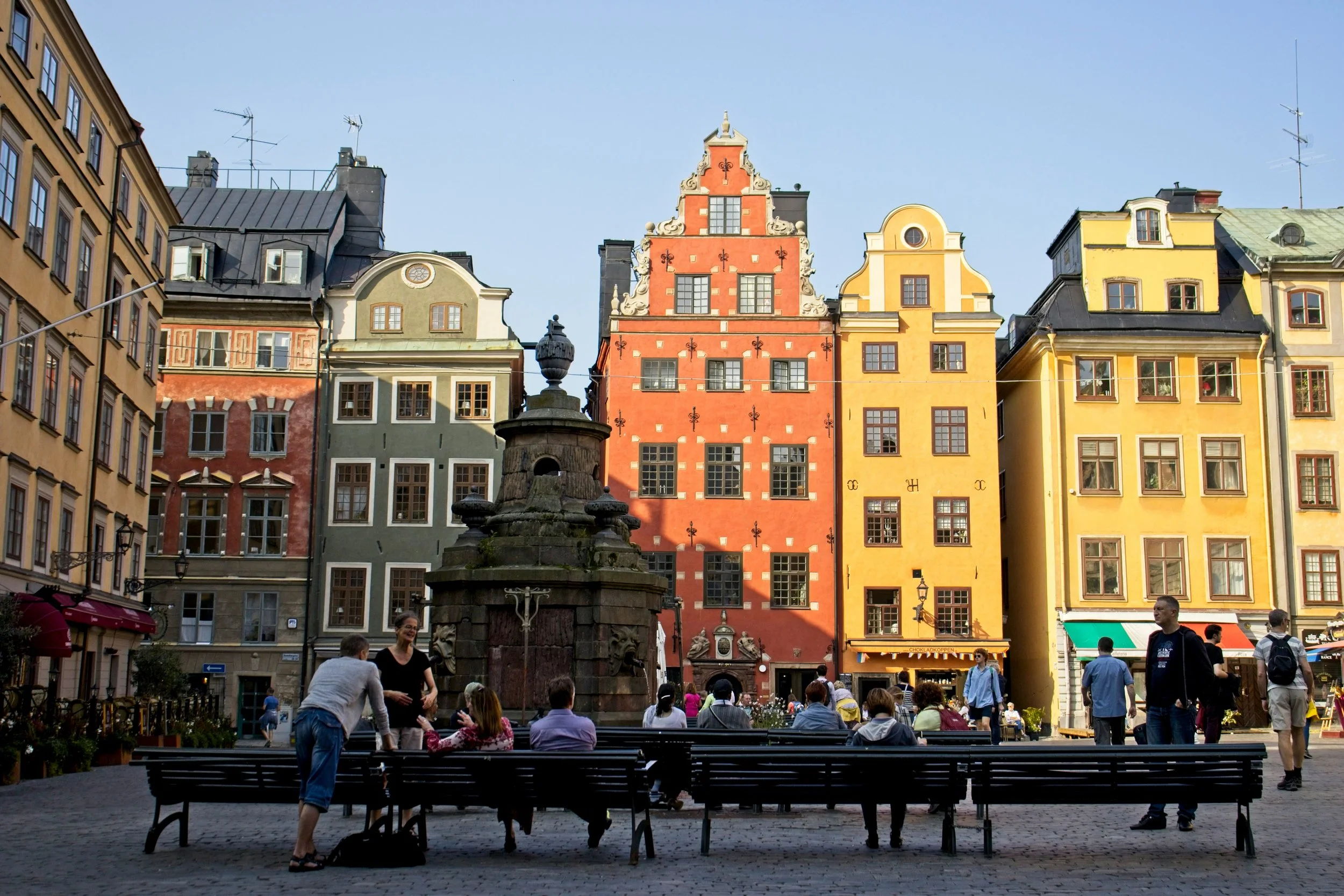 Colorful historic buildings in a European city square with benches and people sitting and talking, a stone fountain in the center, bright blue sky.