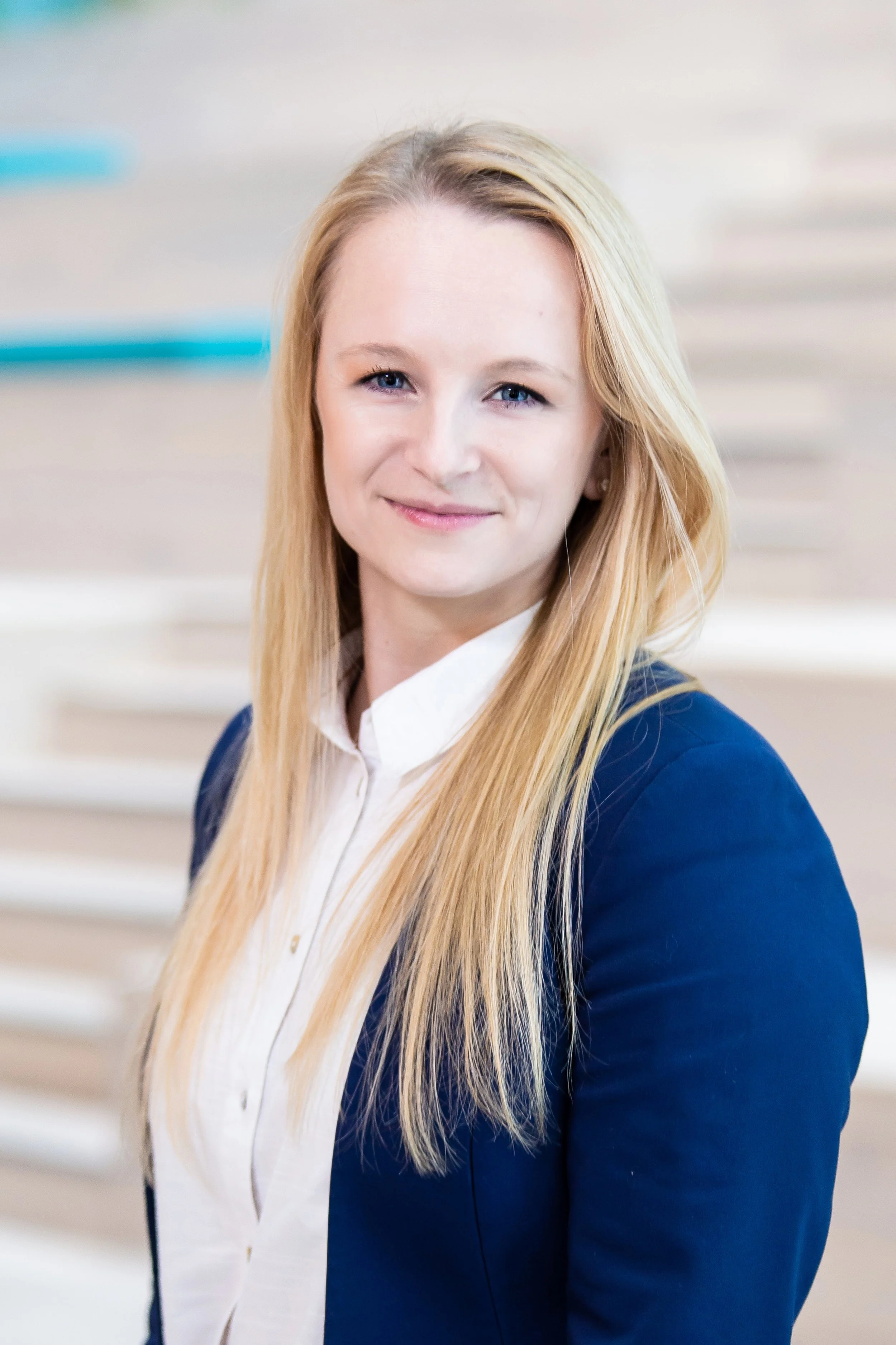 A professional woman with long blonde hair, wearing a navy blazer and white blouse, smiling in an office setting.