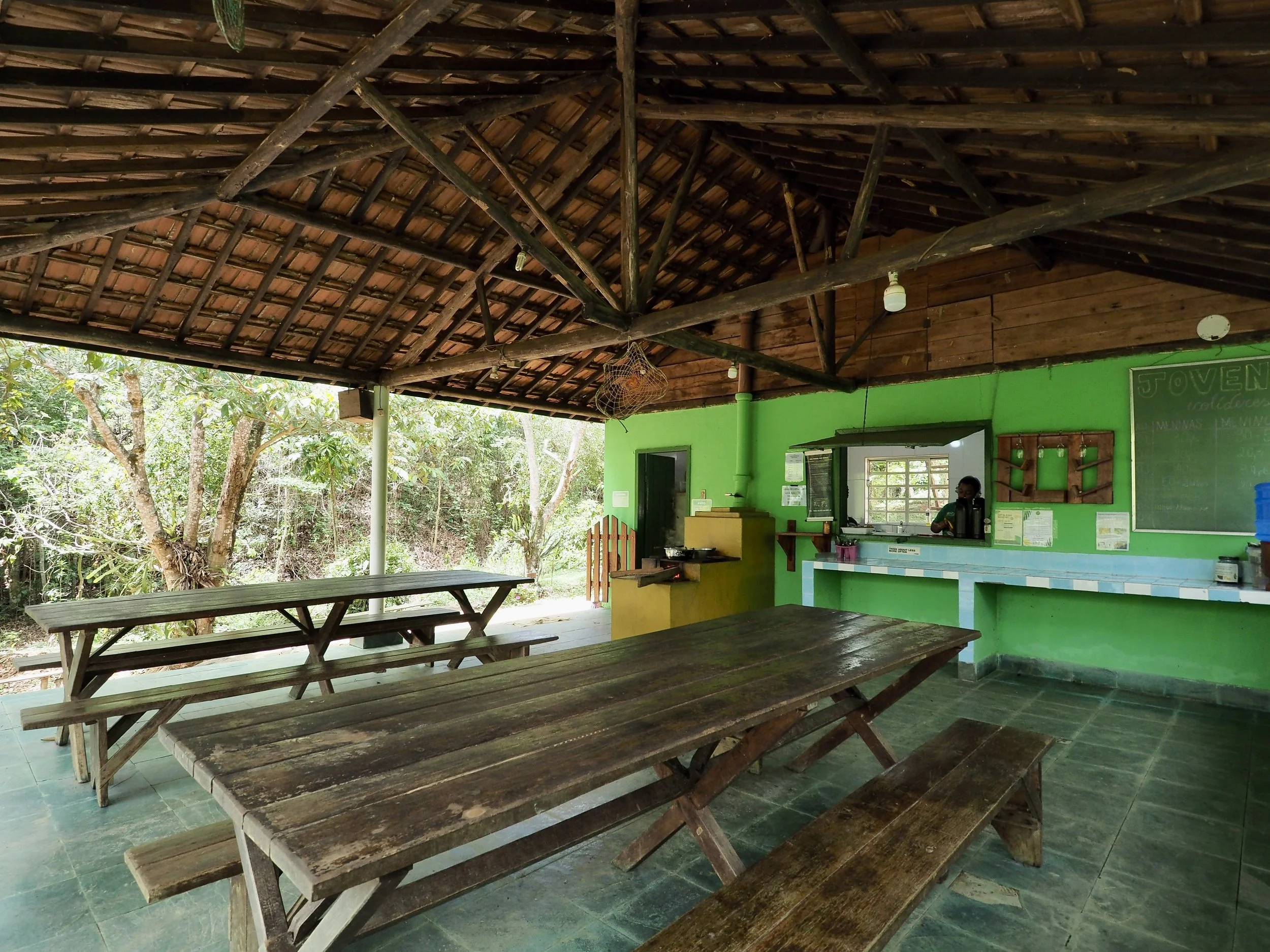 Open-air rustic dining area with green walls, wooden picnic tables, and a food counter with a woman serving drinks. Surrounded by trees, with a tiled floor and a thatched roof.