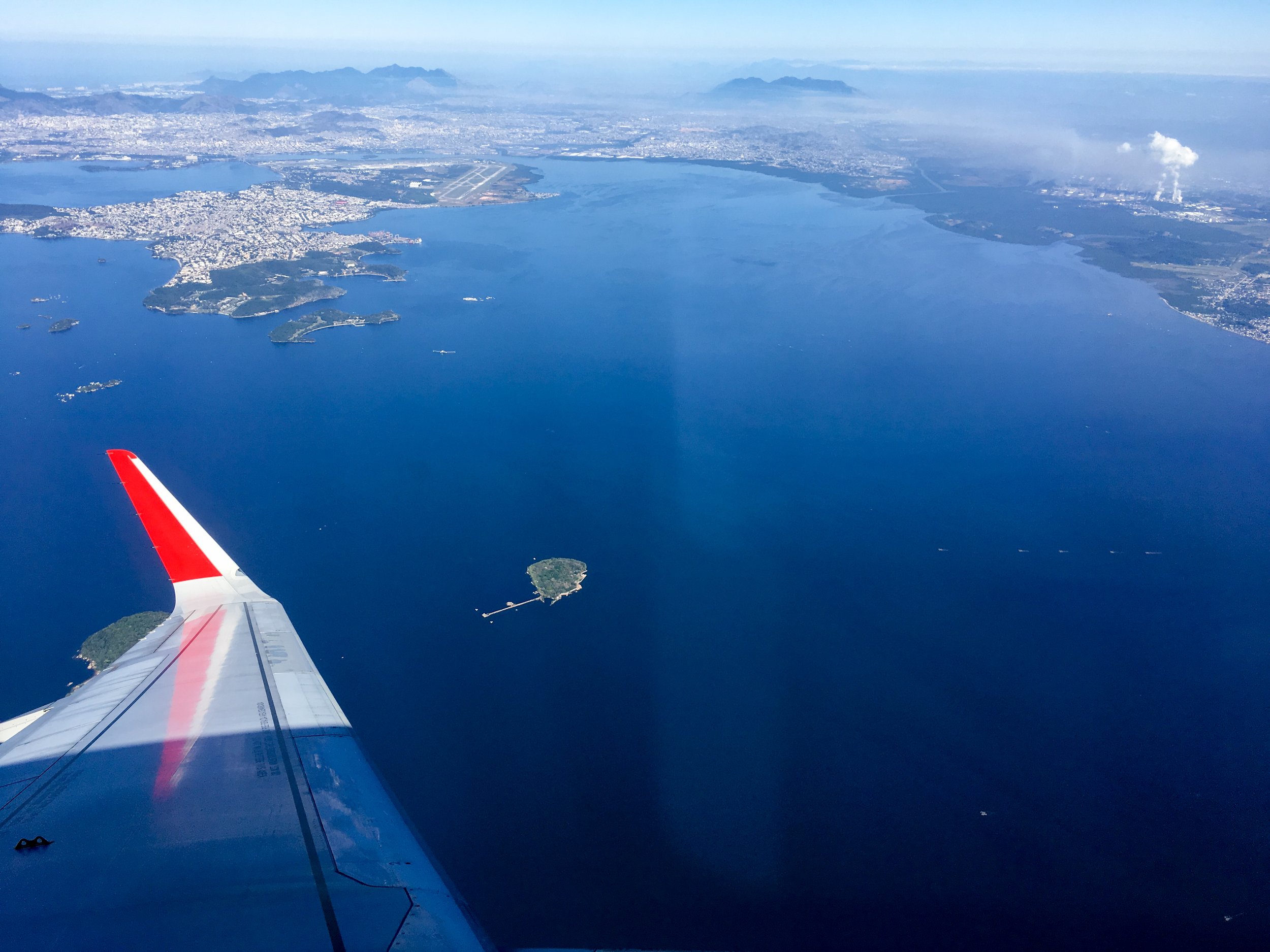 Aerial view from an airplane showing a large blue body of water with small islands, coastal city, and mountains in the distance.