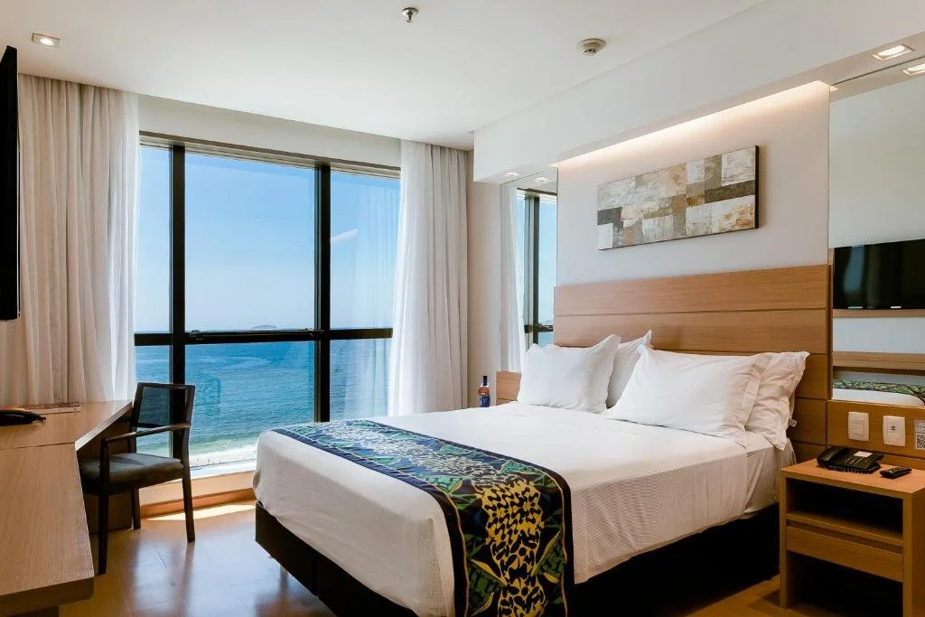 Hotel room with a large window overlooking the ocean, a bed with white sheets and pillows, a wooden headboard, a desk with a chair, and a small TV mounted on the wall.