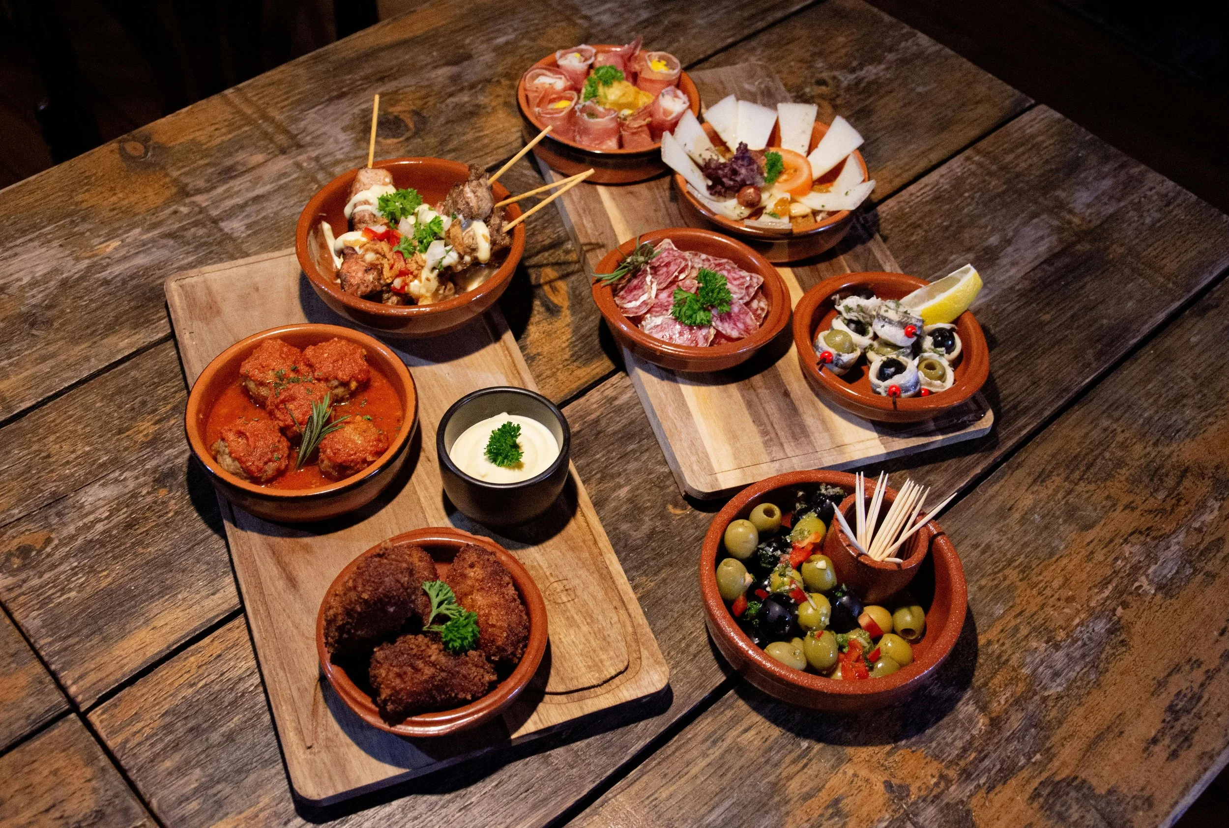 Assorted Mediterranean appetizers including olives, breaded meatballs, sliced meats, cheese, and various dips, all arranged on small terracotta dishes and a wooden serving board on a rustic wooden table.