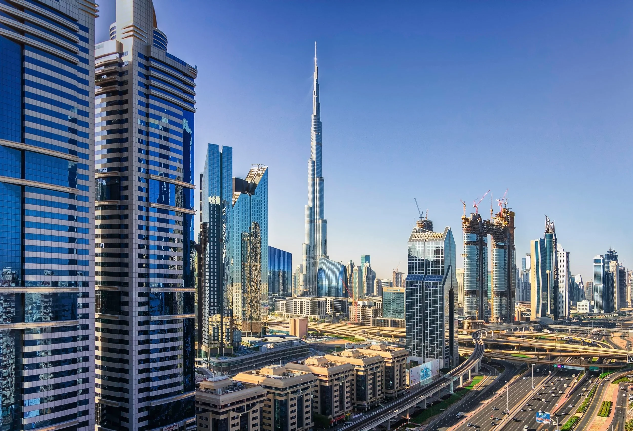 Skyscrapers and high-rise buildings in Dubai, with the Burj Khalifa in the center, under a clear blue sky, with roads and highways in the foreground.